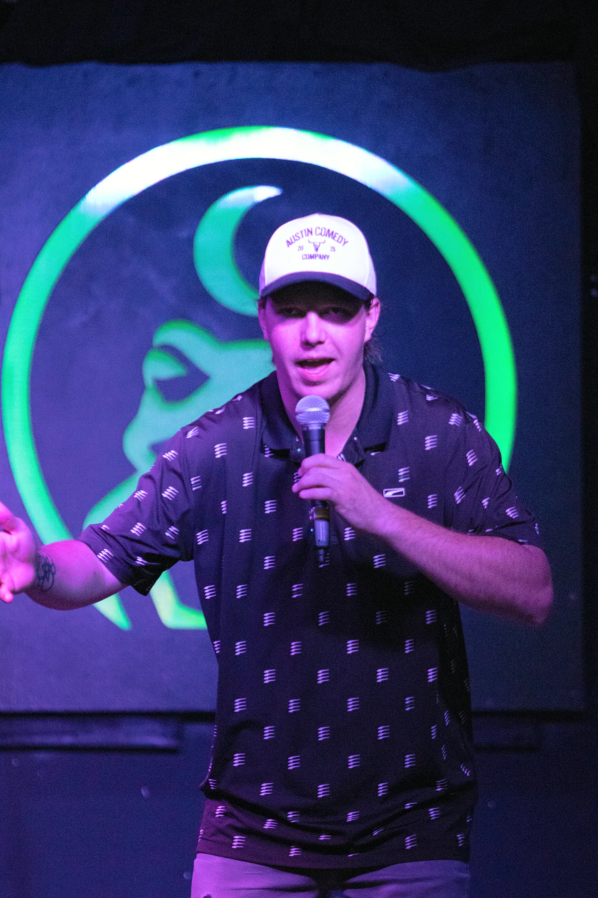 A comic in an Austin Comedy Company cap and a patterned polo, one arm flung out mid-story, lit violet against the green ring behind him.