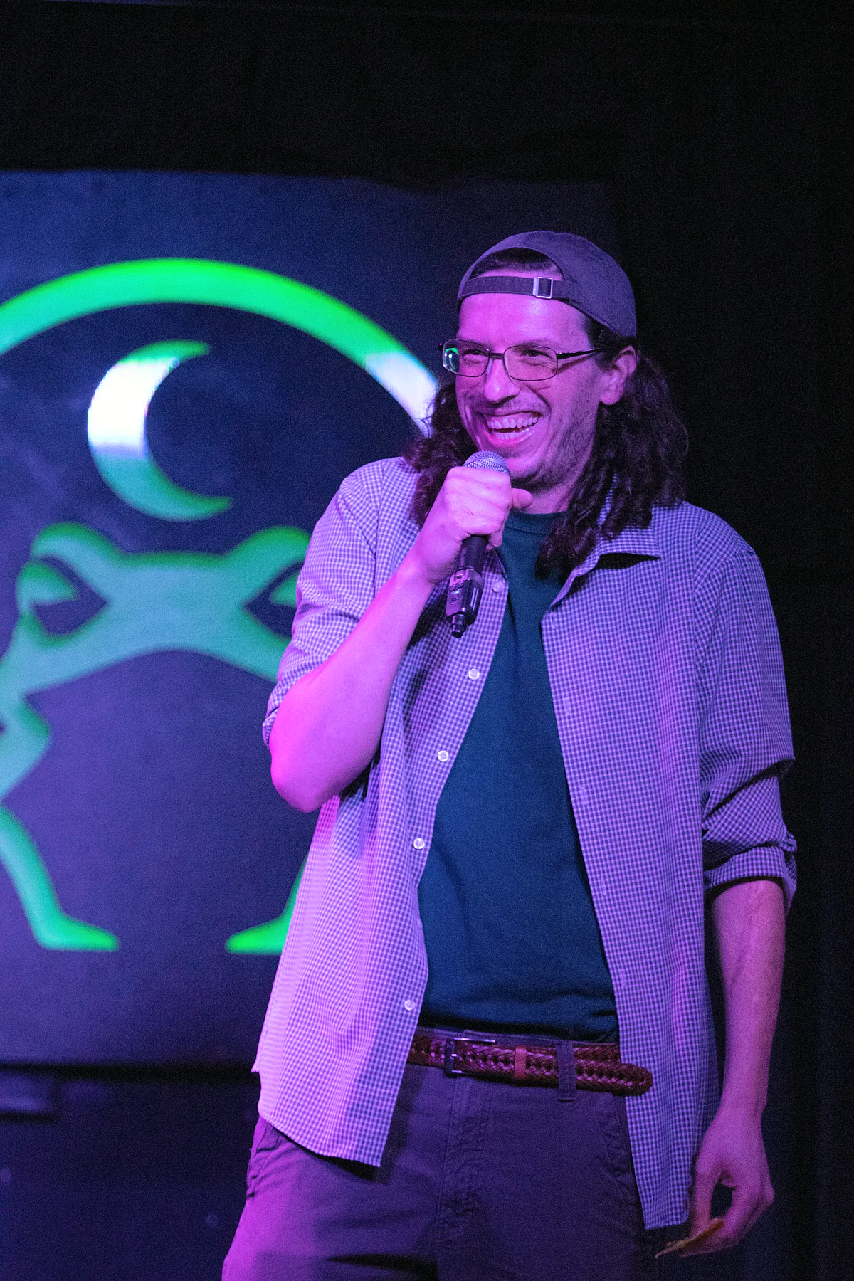 A long-haired comic in a backwards cap and glasses, wearing an open checked shirt over a tee, grinning mid-punchline under magenta light.