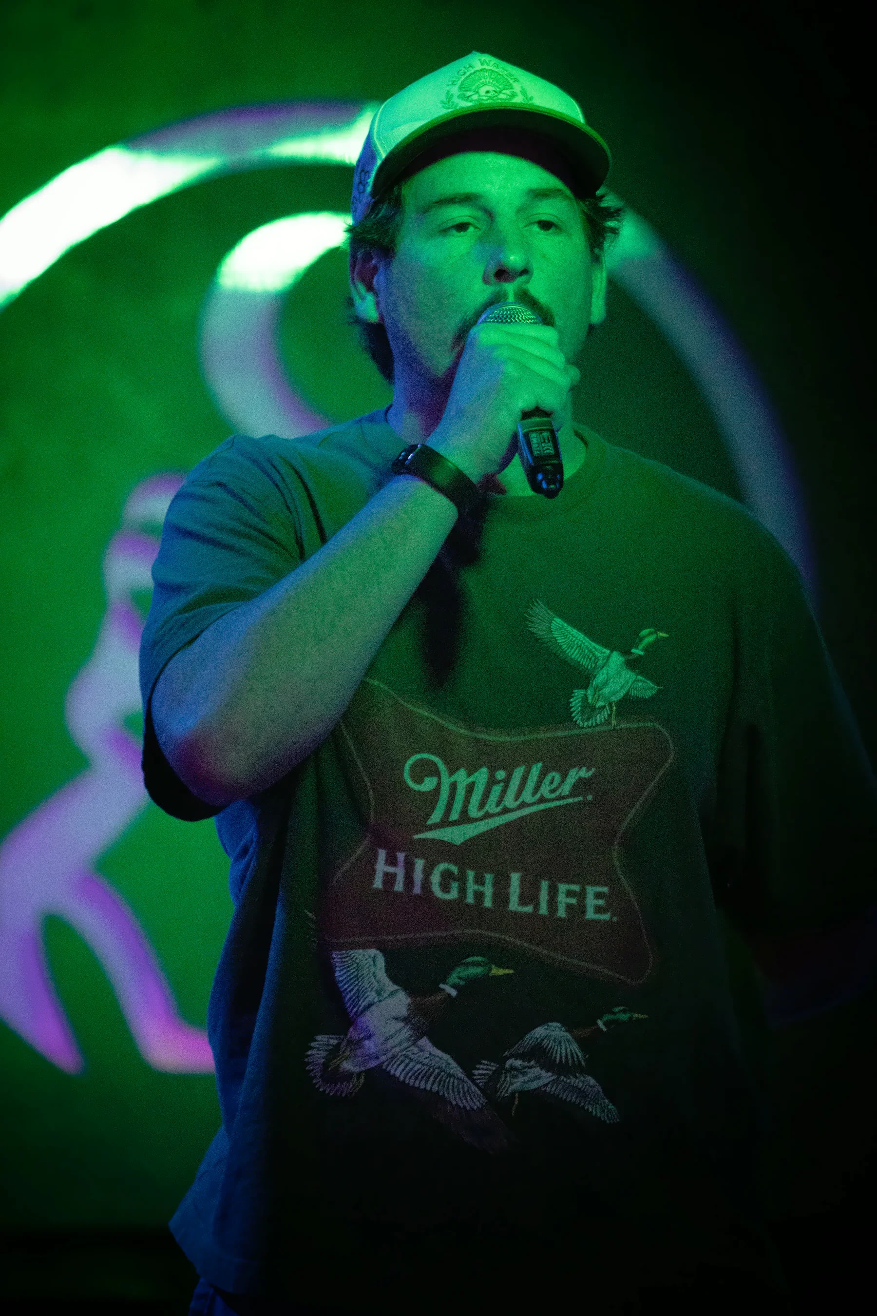 A performer in a light cap and a Miller High Life t-shirt close on the microphone, the lit toad out of focus and half-hidden on the green wall behind.
