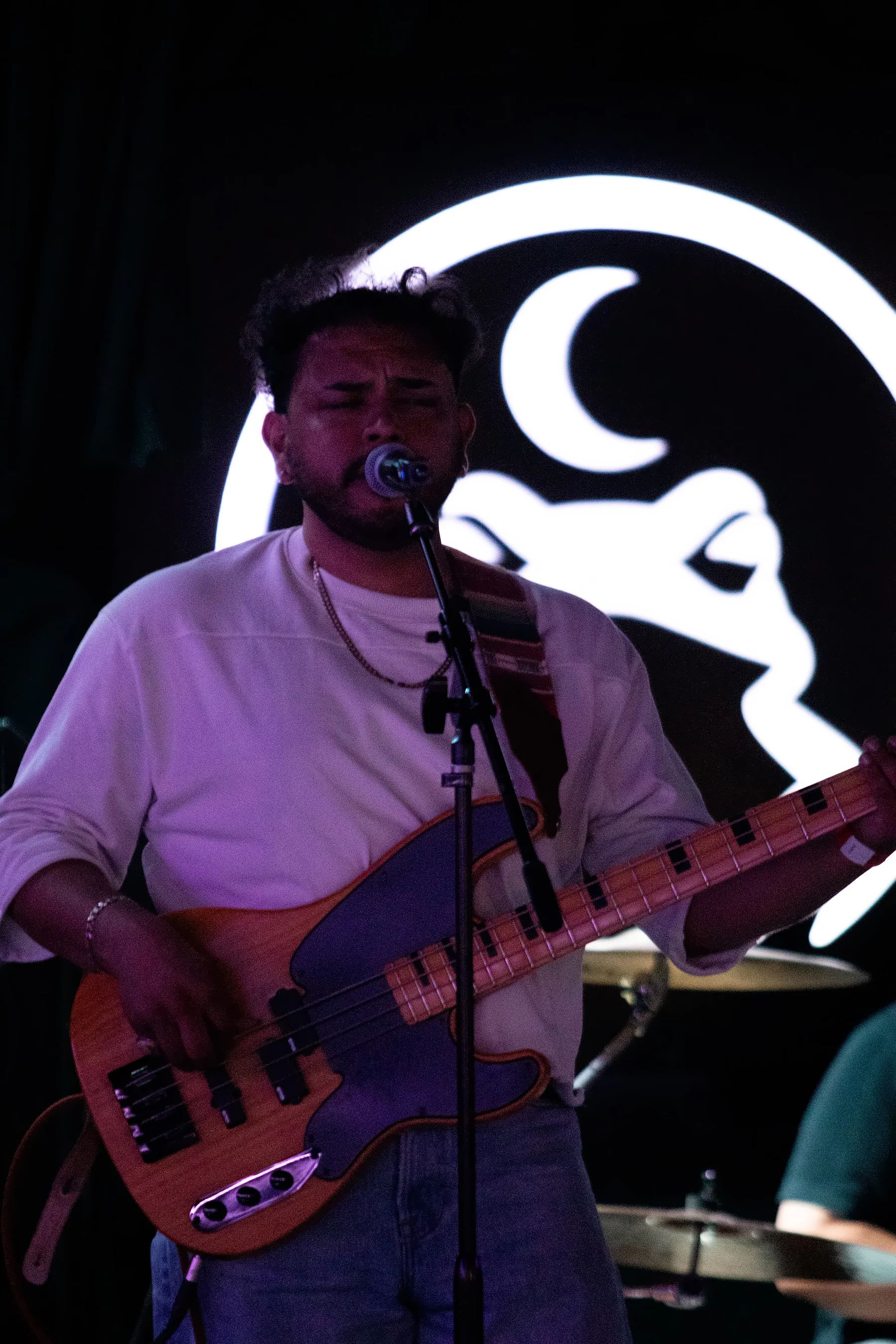 A bassist singing into the microphone in a white t-shirt, the lit toad sign behind his shoulder.