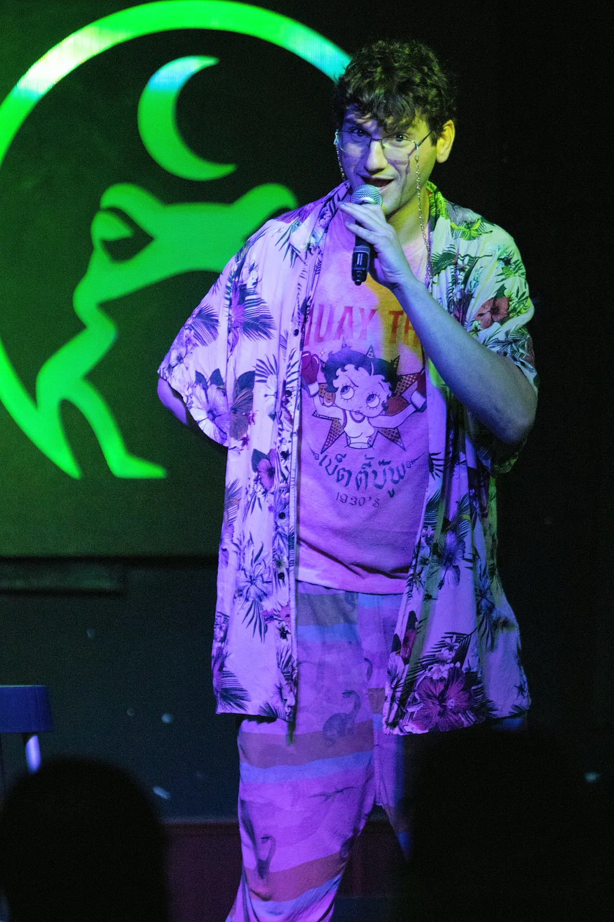 A comic in an open floral shirt over a Betty Boop tee and striped trousers, glasses on a chain, mid-punchline in green light.