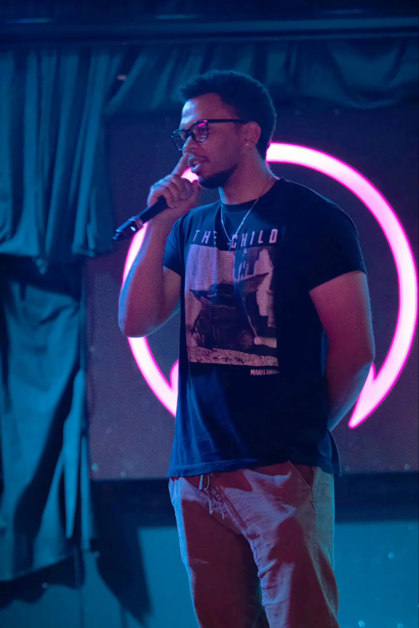 A performer in glasses and a black printed t-shirt with the microphone close to their mouth, the lit toad’s pink ring glowing behind them.