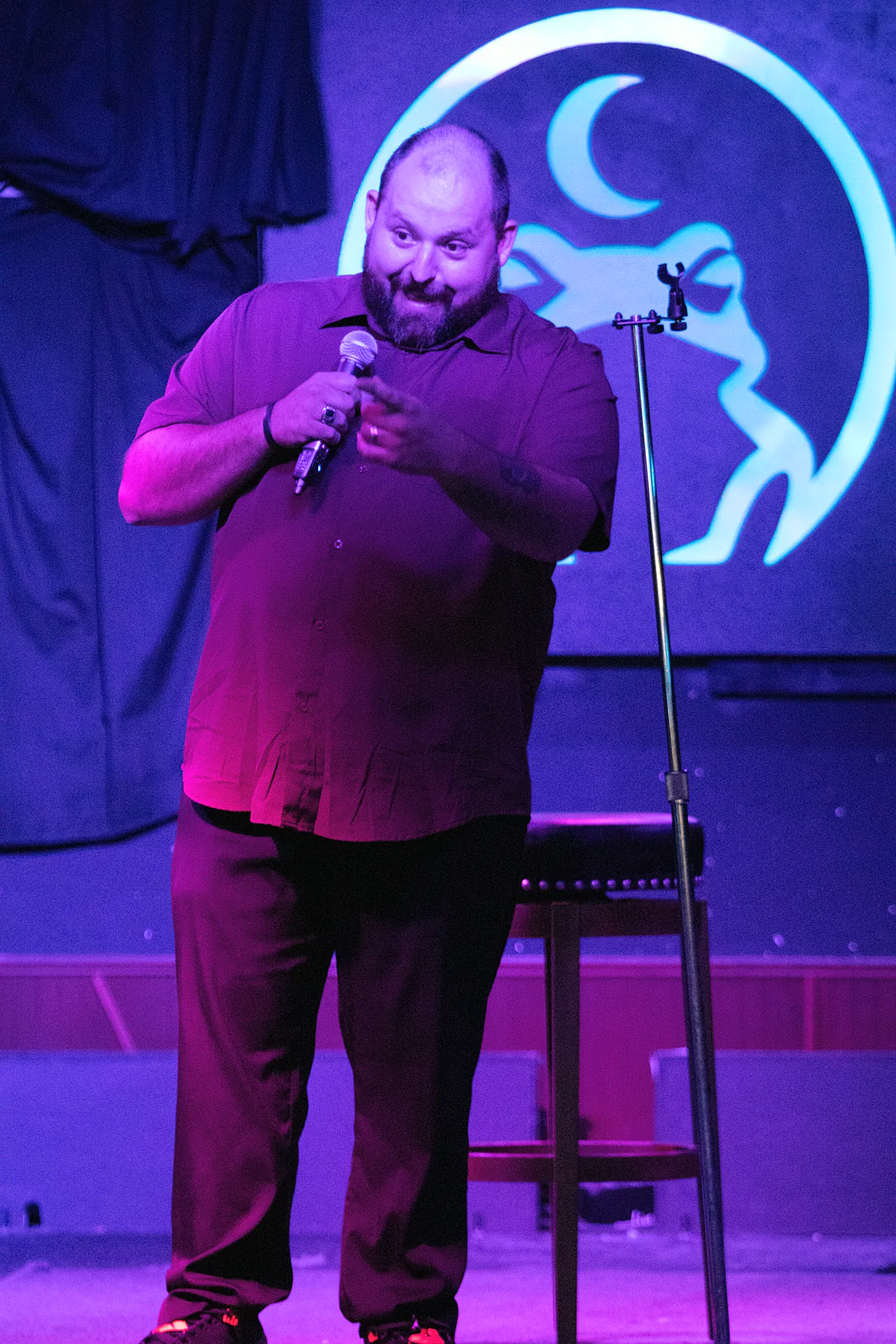 A bearded comic in a burgundy short-sleeved shirt working the microphone with both hands, a mic stand and stool beside him under blue stage light.