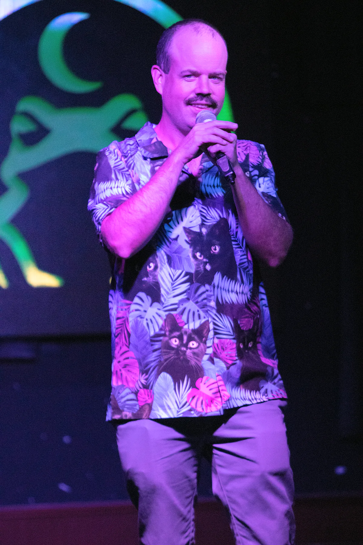 A moustached comic in a tropical shirt printed with cats, both hands on the microphone, smiling out at the room.