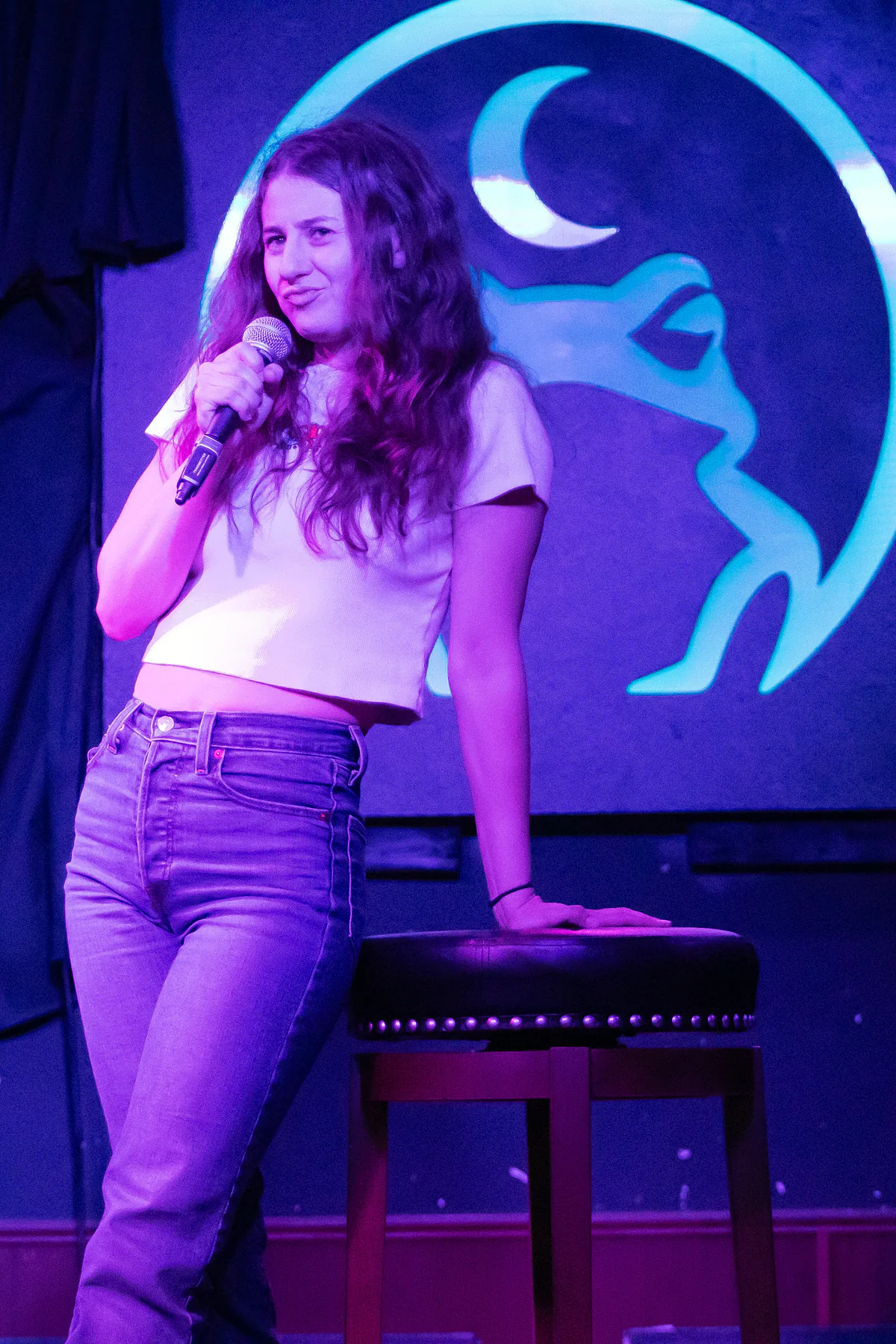 A comic in a white tee and jeans mid-set, one hand on the stool beside her, lit blue and violet.