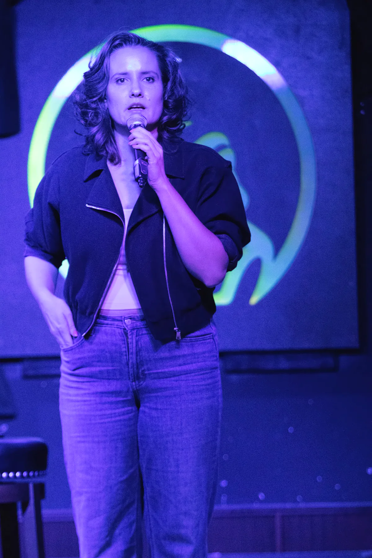 A performer in a dark cropped jacket and jeans with one hand in a pocket and the microphone raised, the lit toad’s green ring glowing behind them.