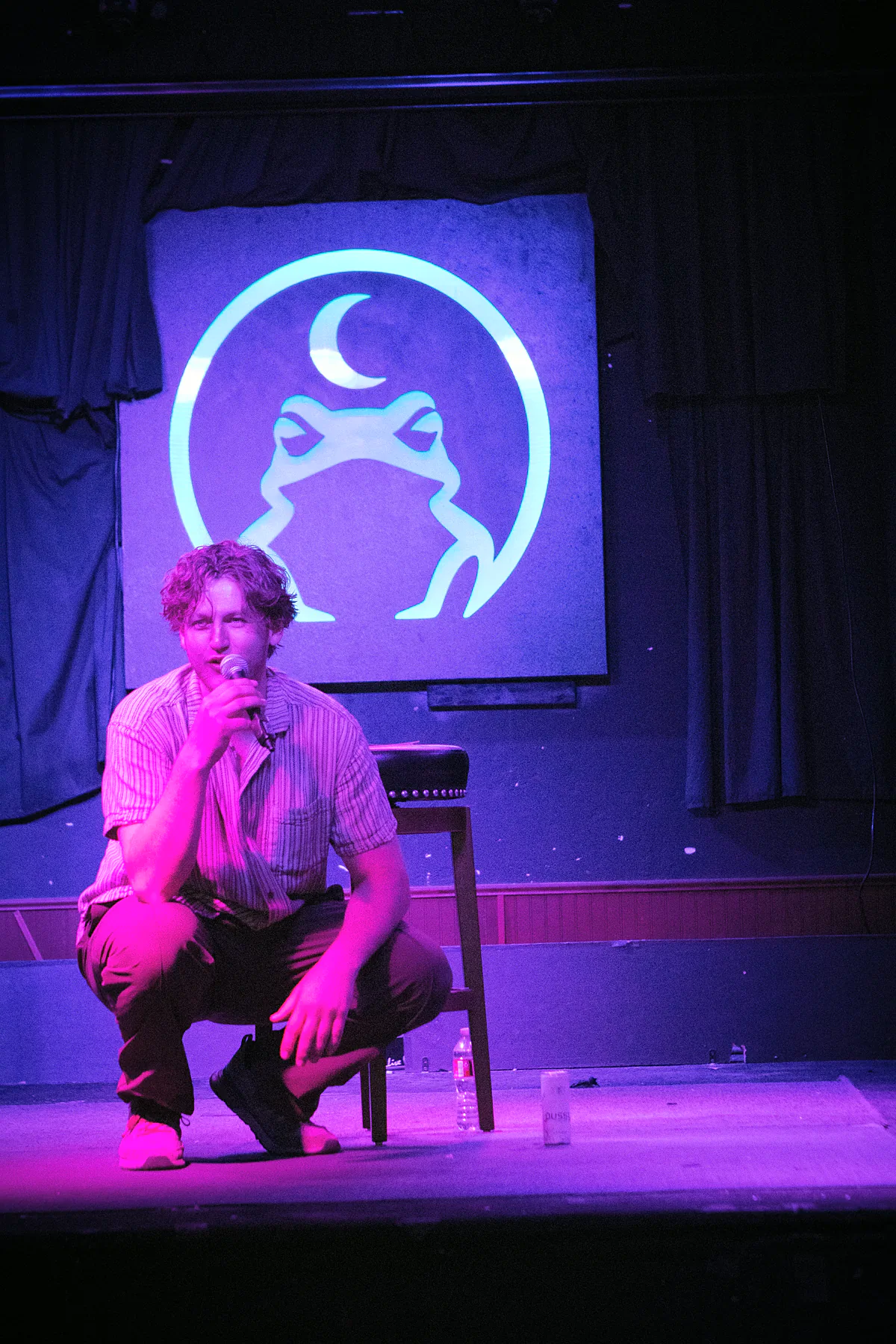 A comic in a striped shirt crouched down at the front of the stage with the microphone, a water bottle and drink by the stool behind him.