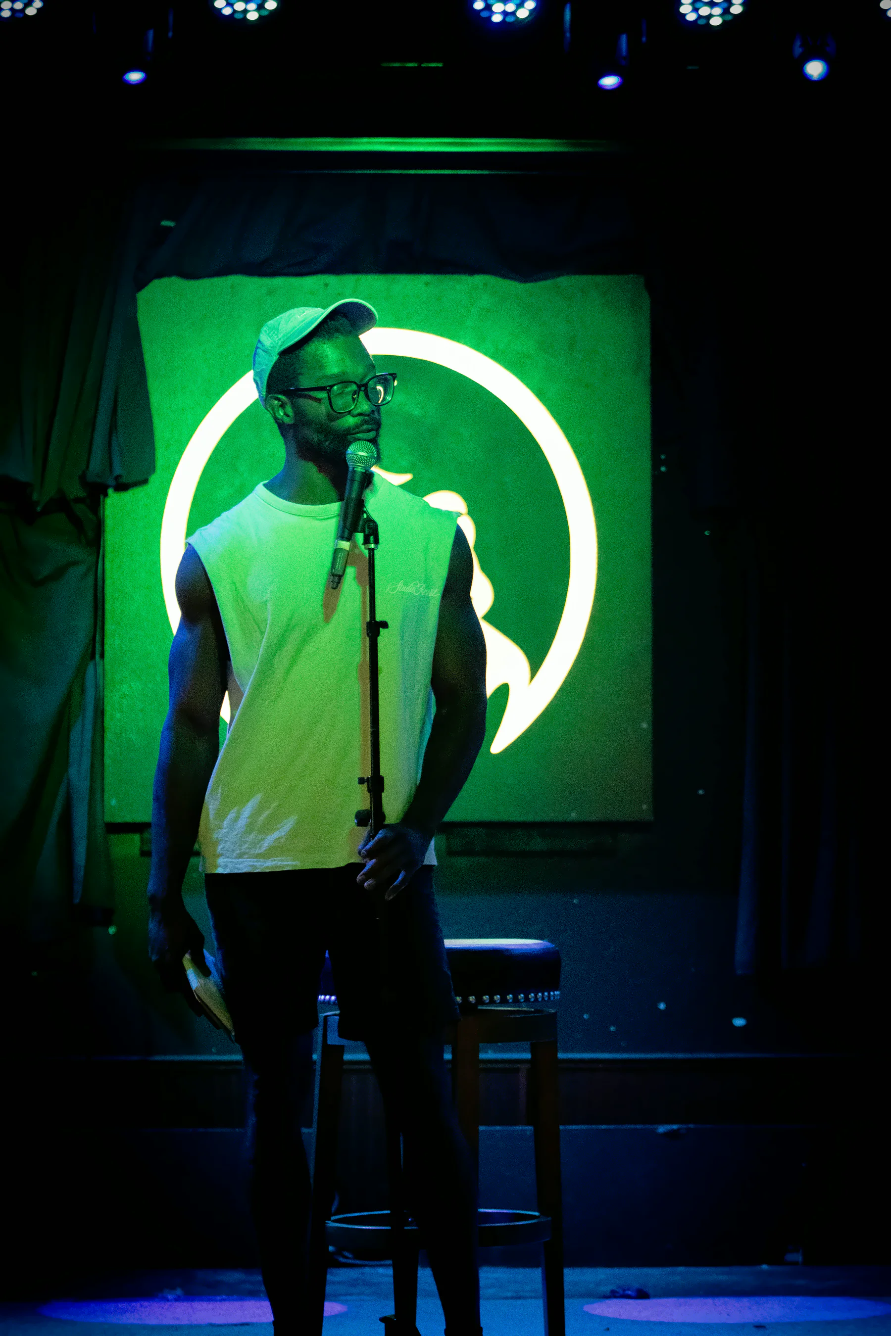 A performer in a pale cap and white sleeveless shirt at the mic stand, the lit toad’s ring glowing around them and their body hiding most of the frog.