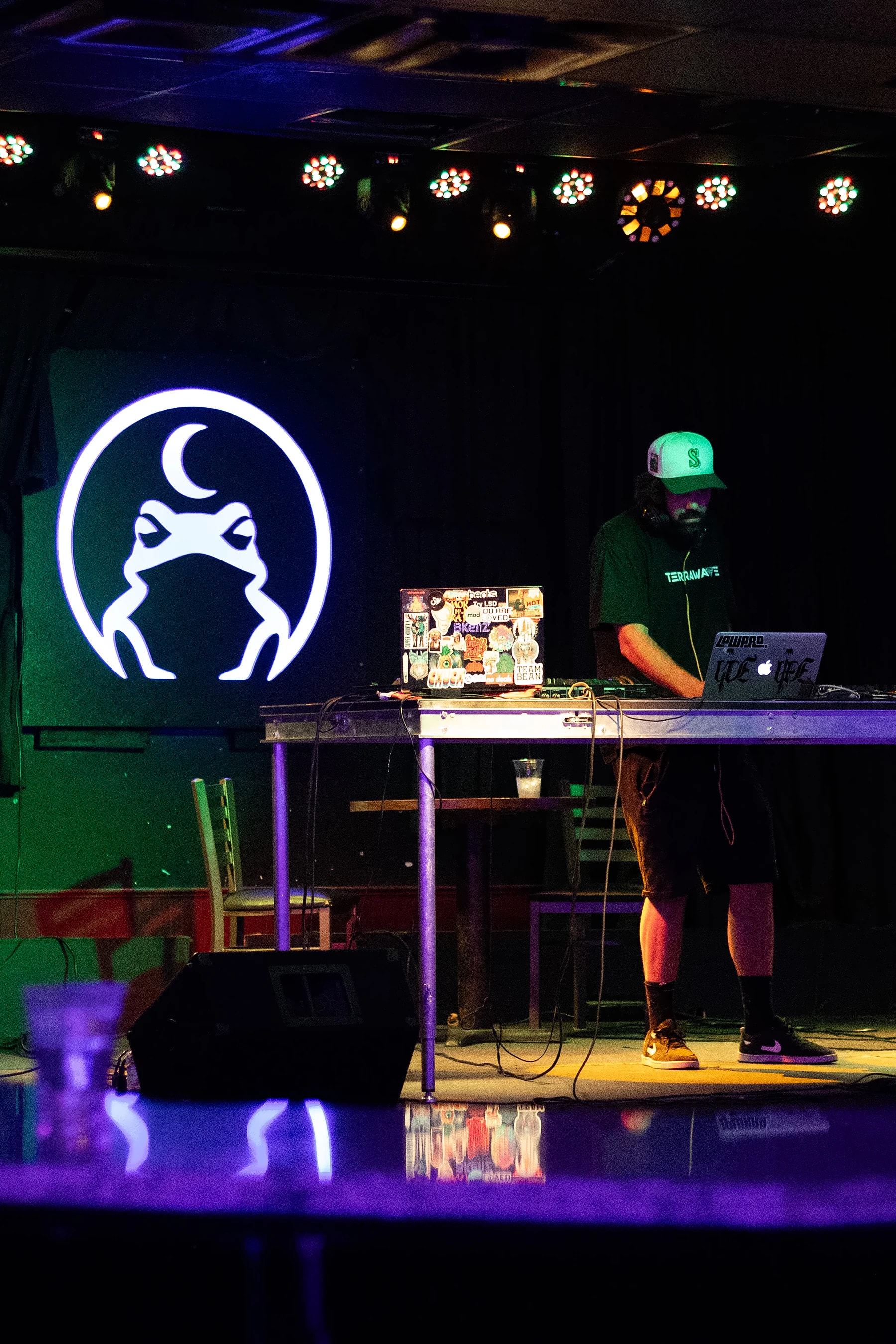 A DJ behind a laptop and controller on the Toad stage, the toad sign lit white on the wall behind.