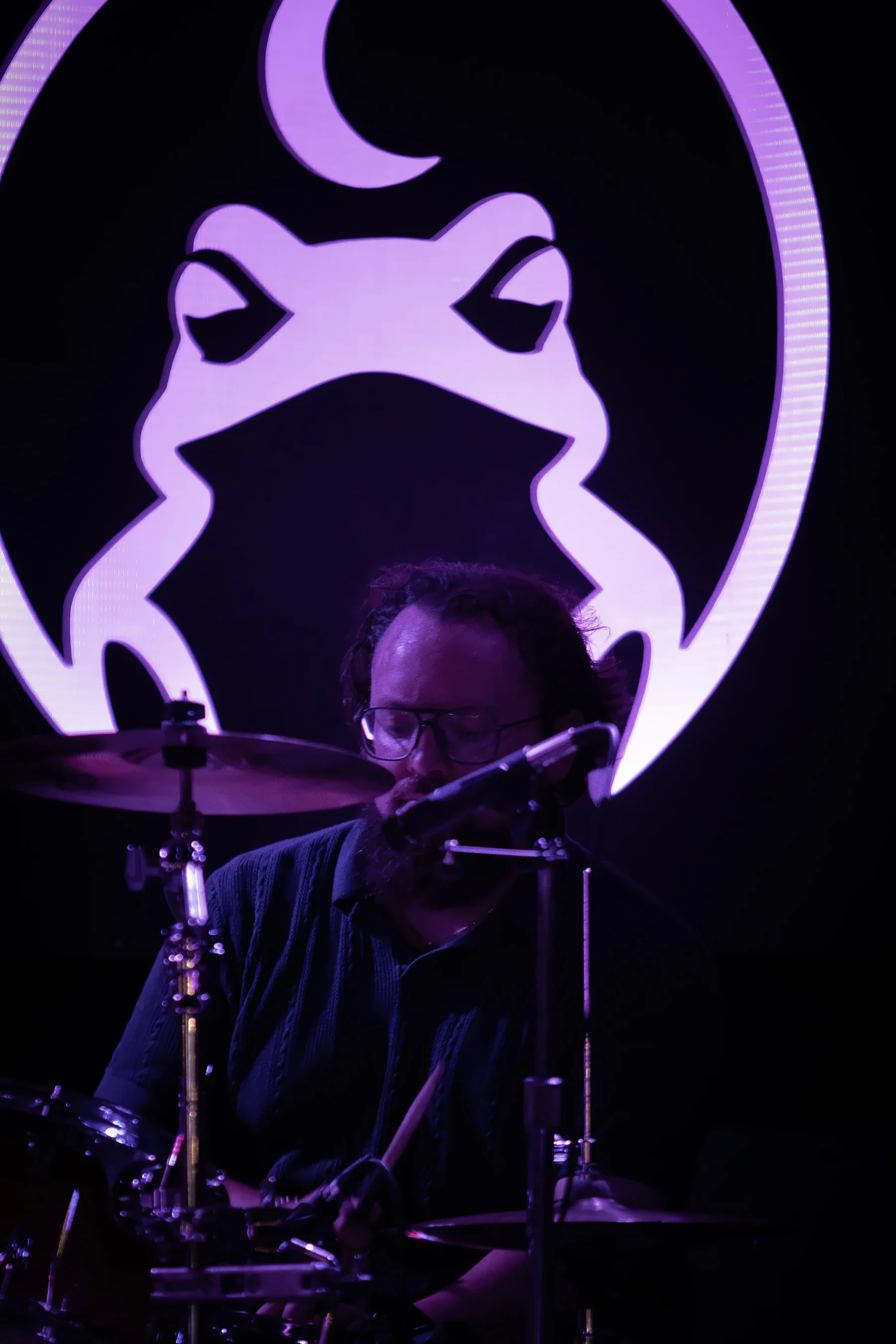 A drummer at the kit behind a vocal mic, the toad sign washed purple and filling the wall above him.