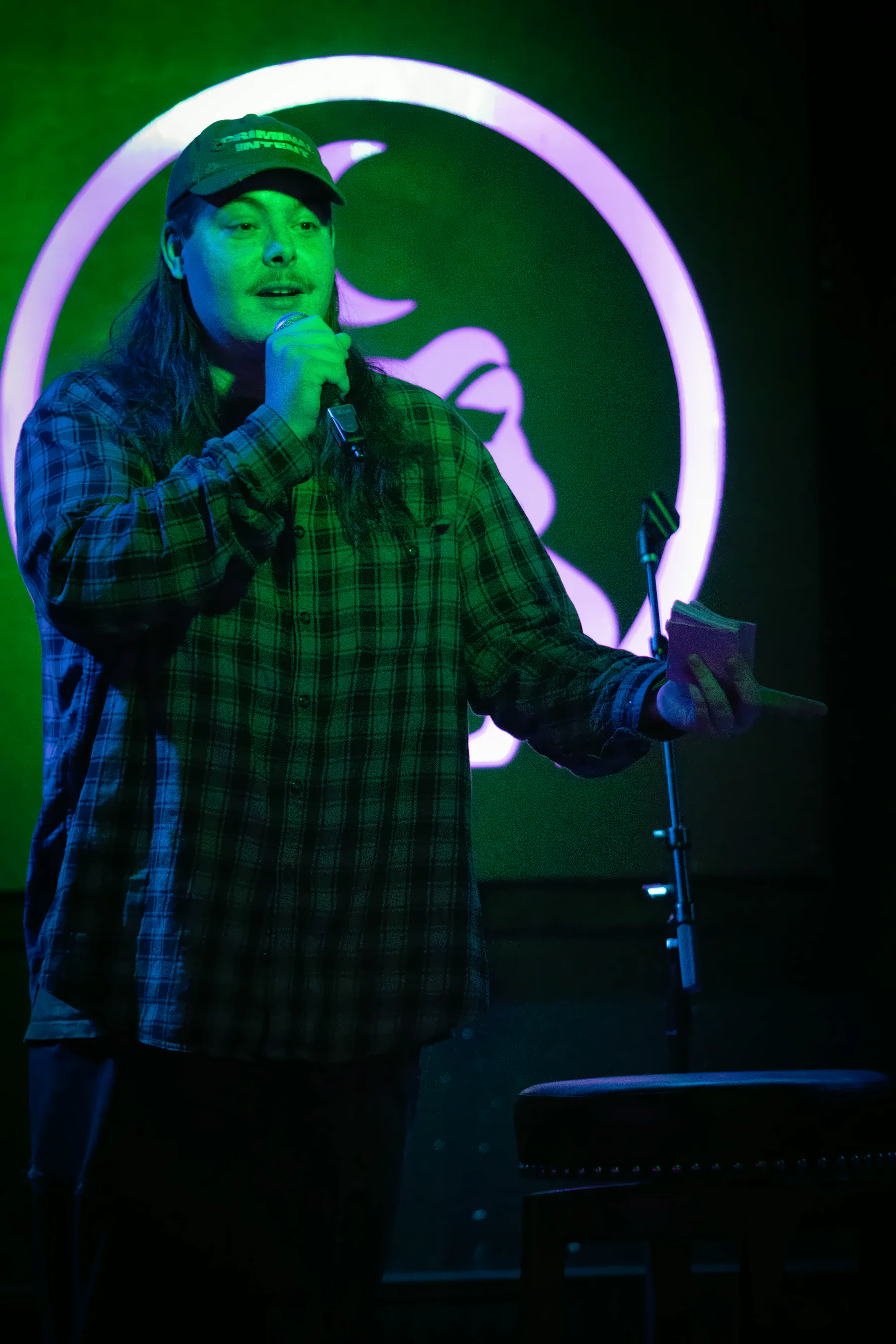 A performer in a baseball cap and plaid flannel with a stack of cards in one hand, the lit toad glowing pink on the wall behind them.