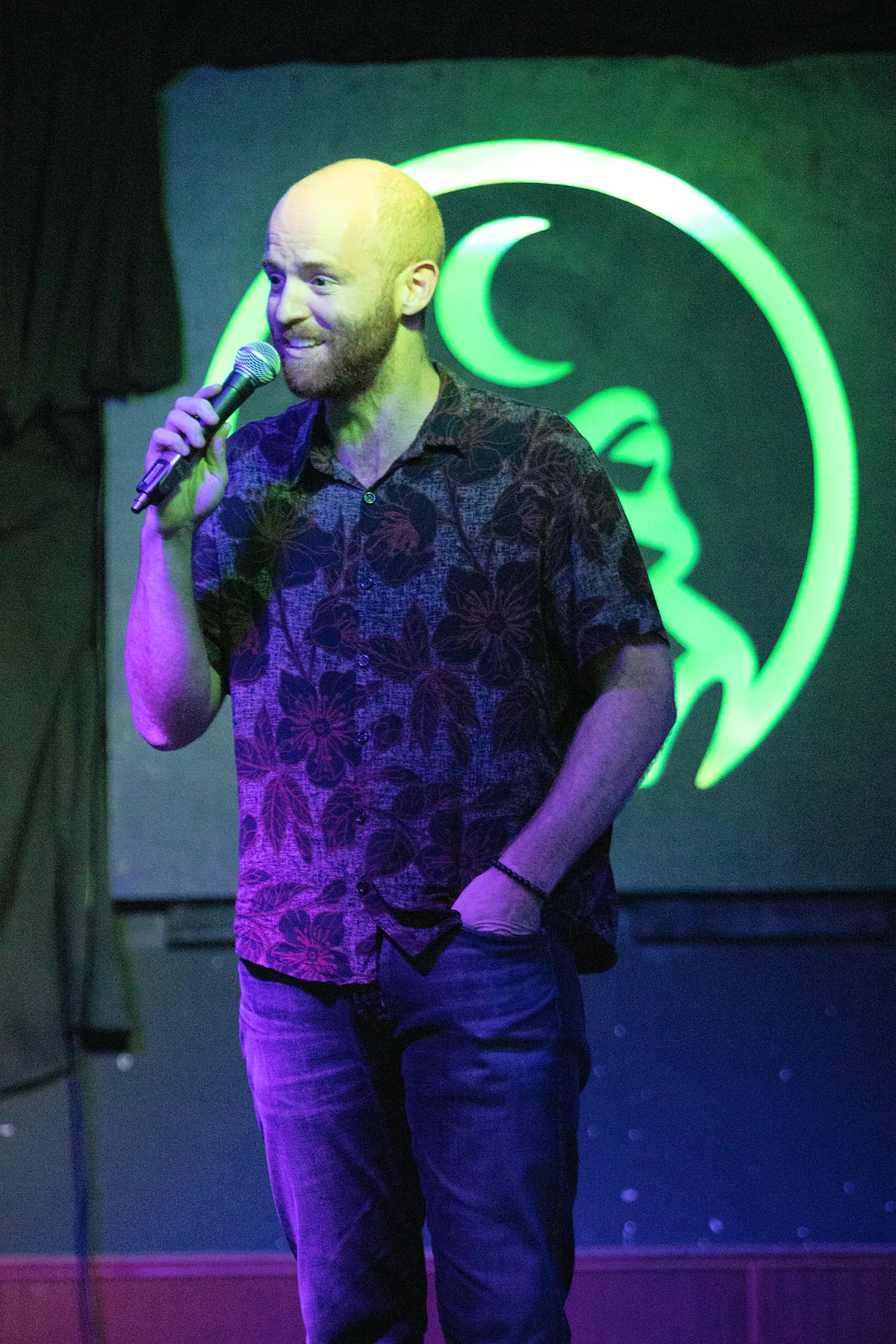 A bearded comic in a dark floral shirt, one hand in his pocket and the other on the microphone, grinning under green light.