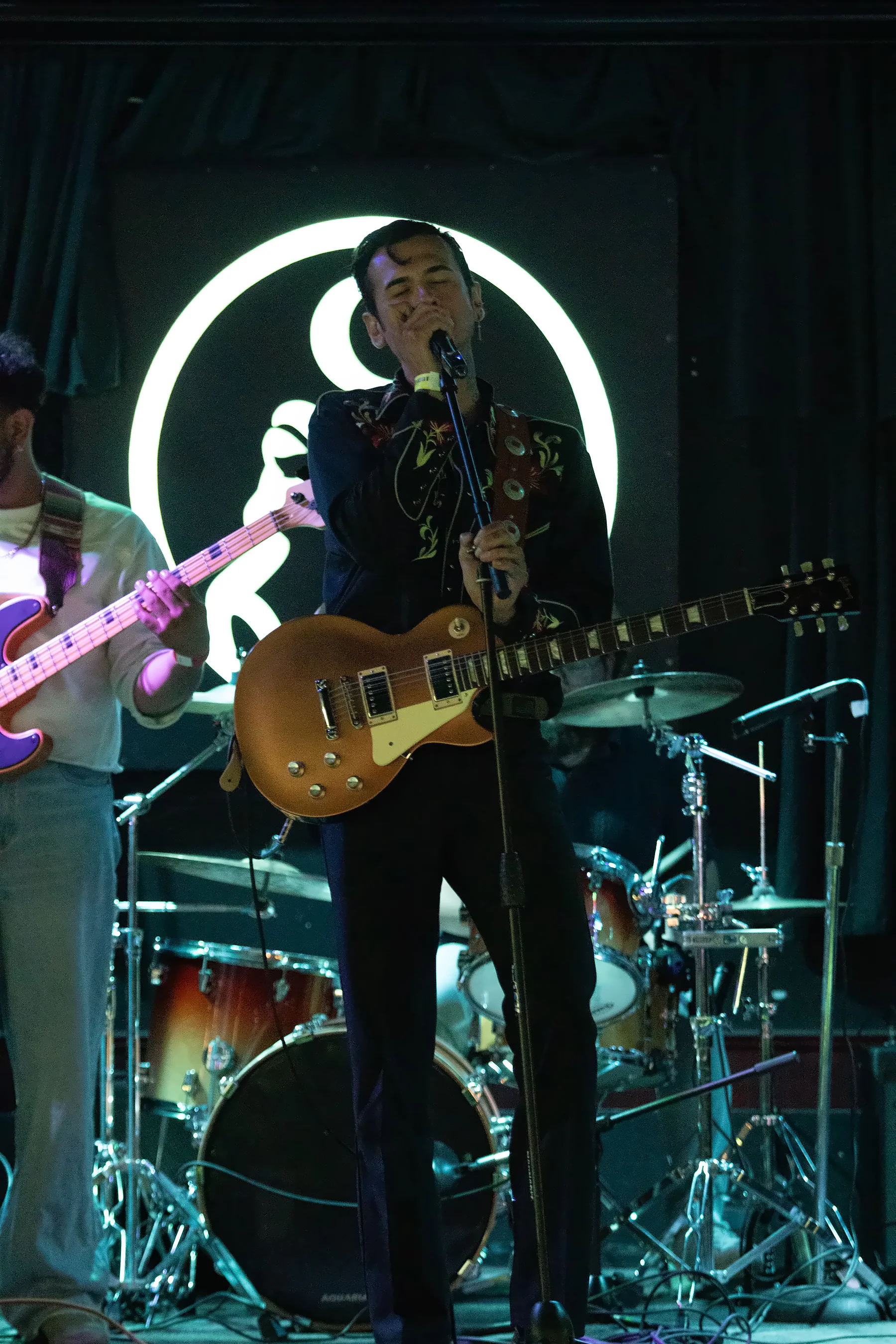 A singer in an embroidered western jacket at the microphone with a gold-top Les Paul, the lit toad sign glowing white behind him and a bassist and drum kit either side.