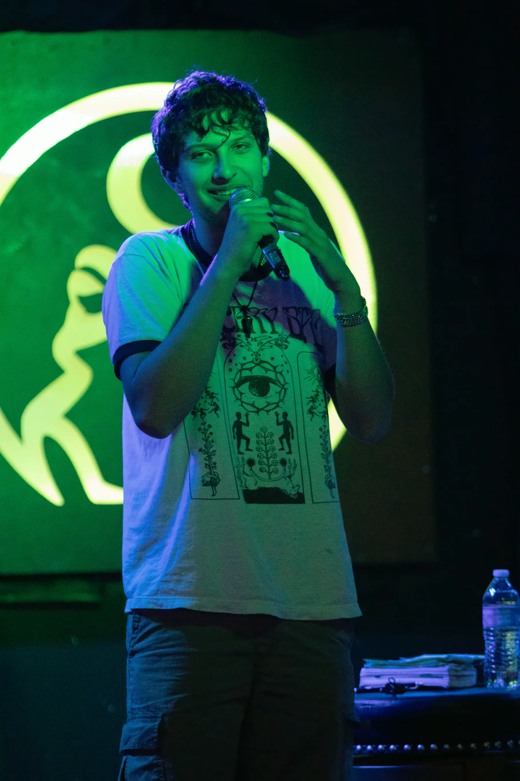 A performer in a ringer t-shirt printed with a large eye, microphone in one hand and the other raised open beside it, the lit toad glowing green behind.