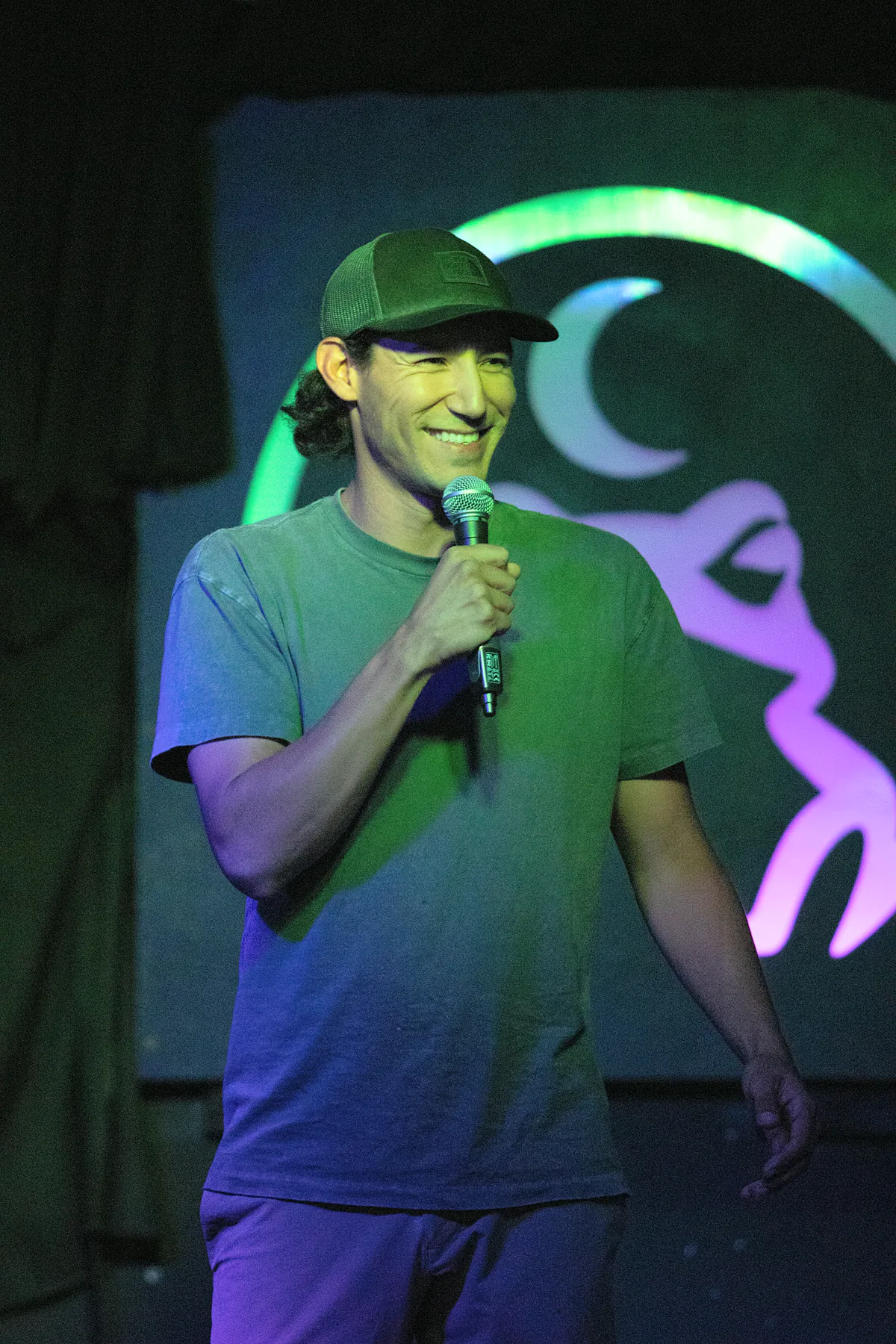 A comic in a cap and grey tee laughing mid-set, caught between green and violet stage light.