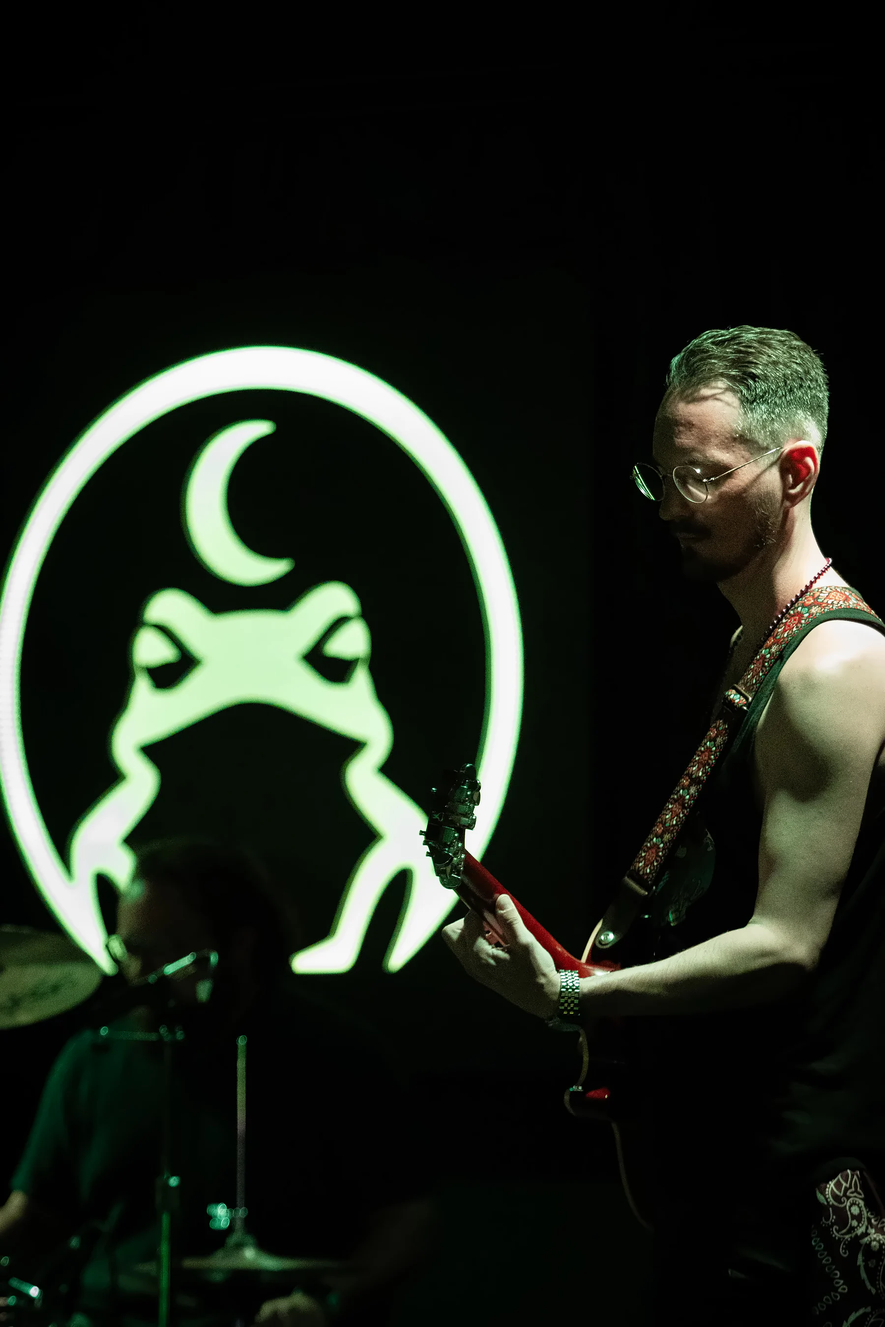 A guitarist in sunglasses and a tank top playing a red guitar, the toad sign lit bright green on the wall beside him.