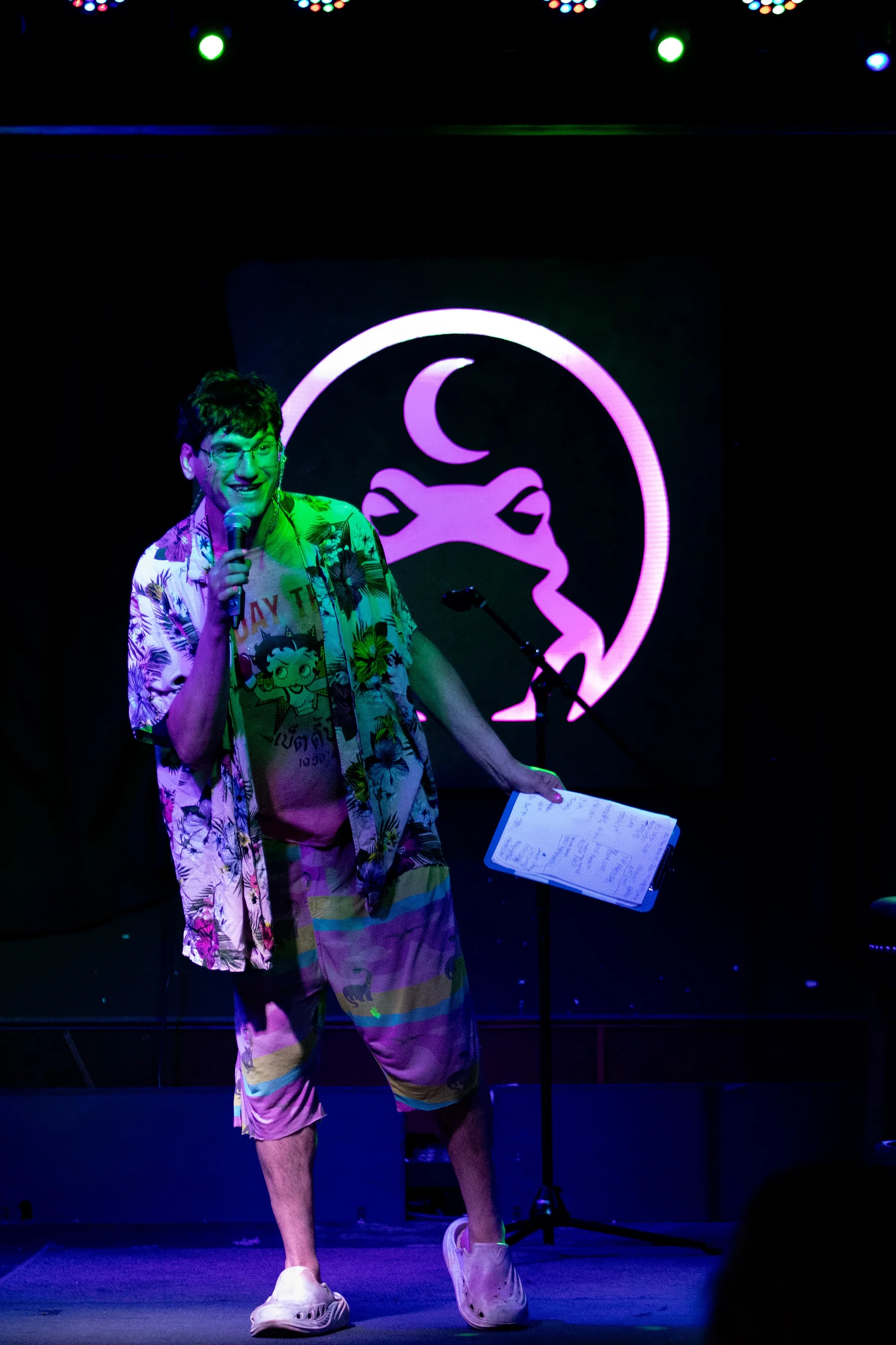 A comic in a floral shirt performing with a clipboard of notes, the lit toad glowing pink behind him.