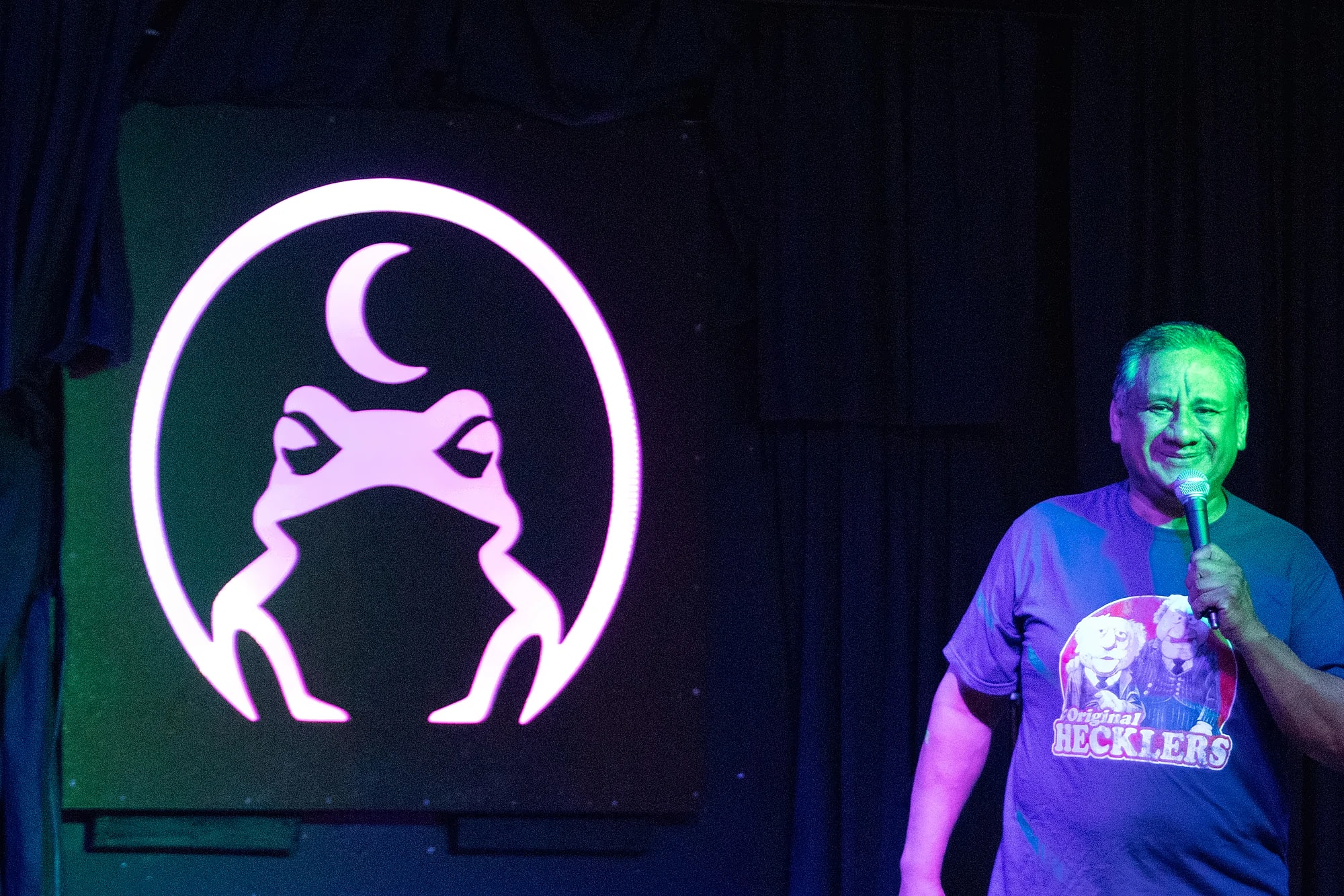 A comic on stage at The Midnight Toad, microphone in hand, standing beside the venue’s lit toad sign.