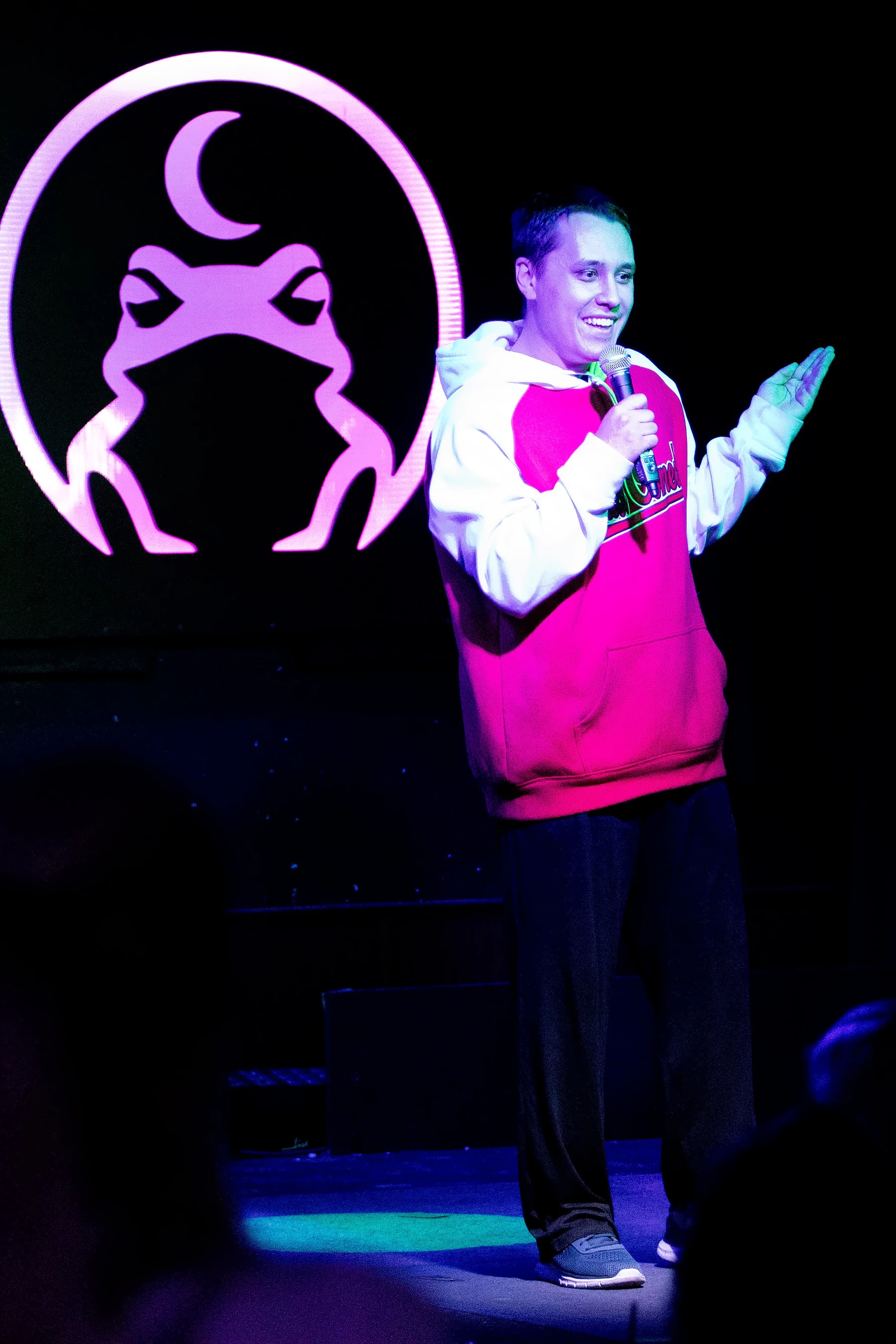 A comic in a red and white hoodie gesturing on stage beside the lit toad.