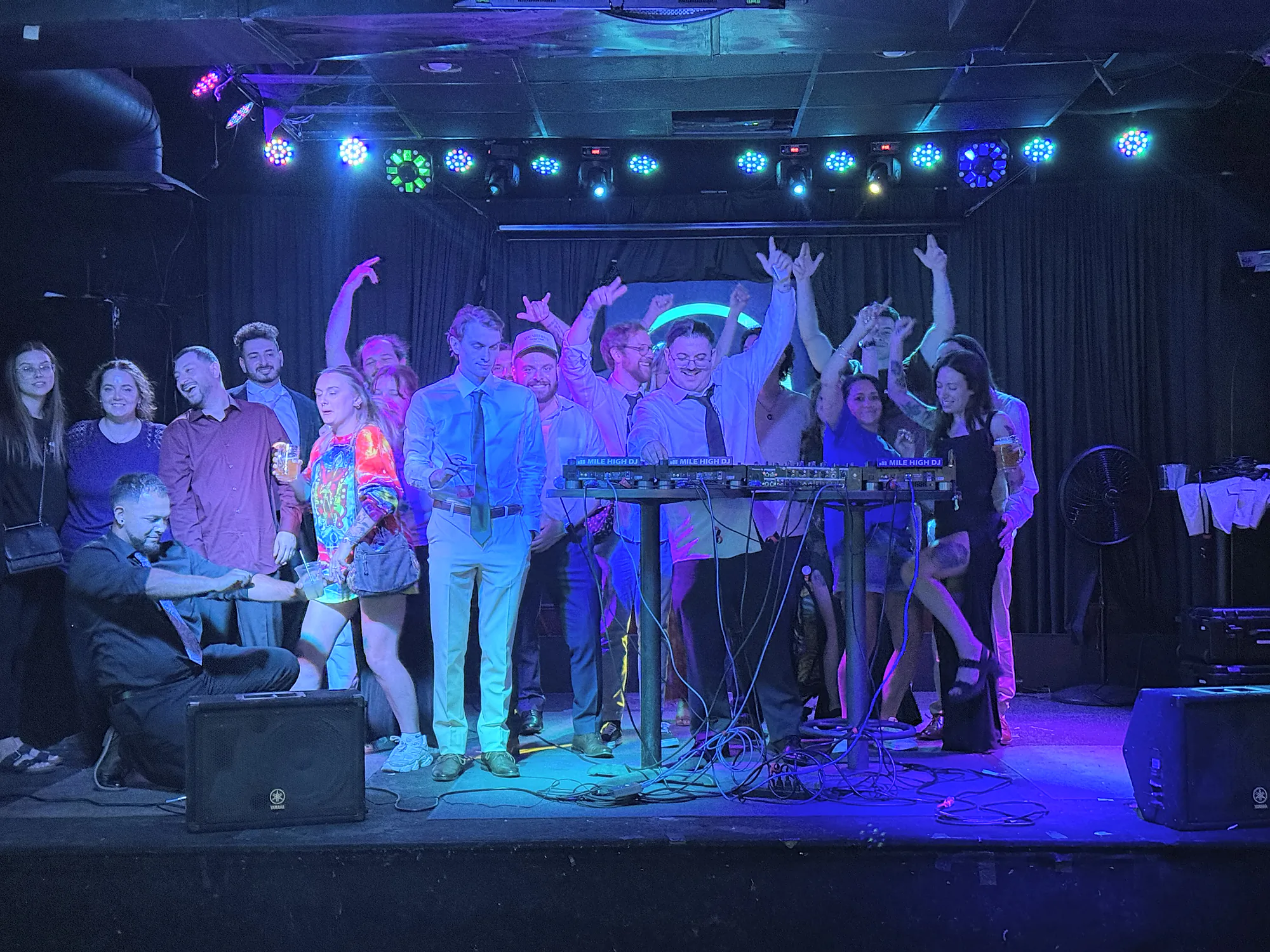 A whole room’s worth of people crowded onto the stage behind a keyboard rig, arms thrown up, under the full colour wash of the light bar.