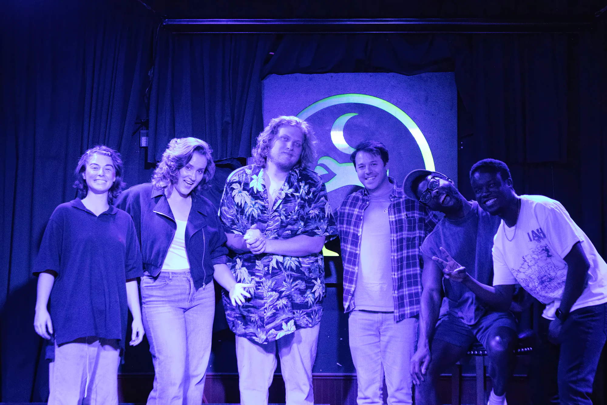Six performers lined up together on stage under blue light, the lit toad’s green ring and crescent glowing above their heads.