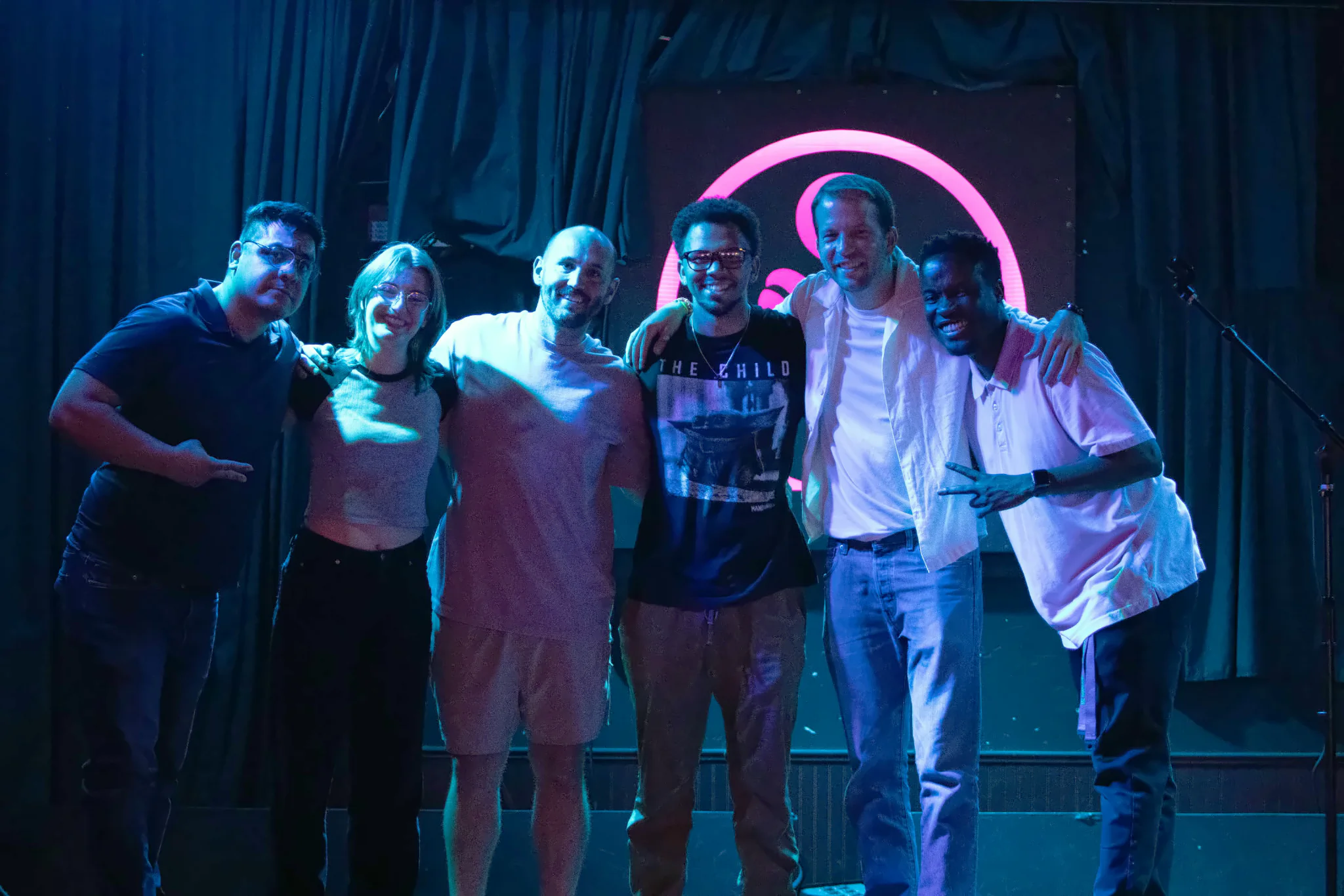 Six performers posed together on stage with arms around each other, the lit toad’s pink ring glowing between their heads.