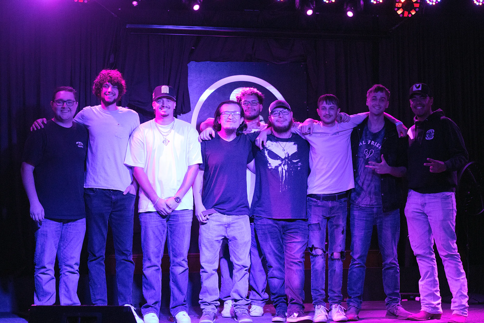 Nine performers lined up across the stage after the show, arms around each other, a couple throwing horns, under purple wash.