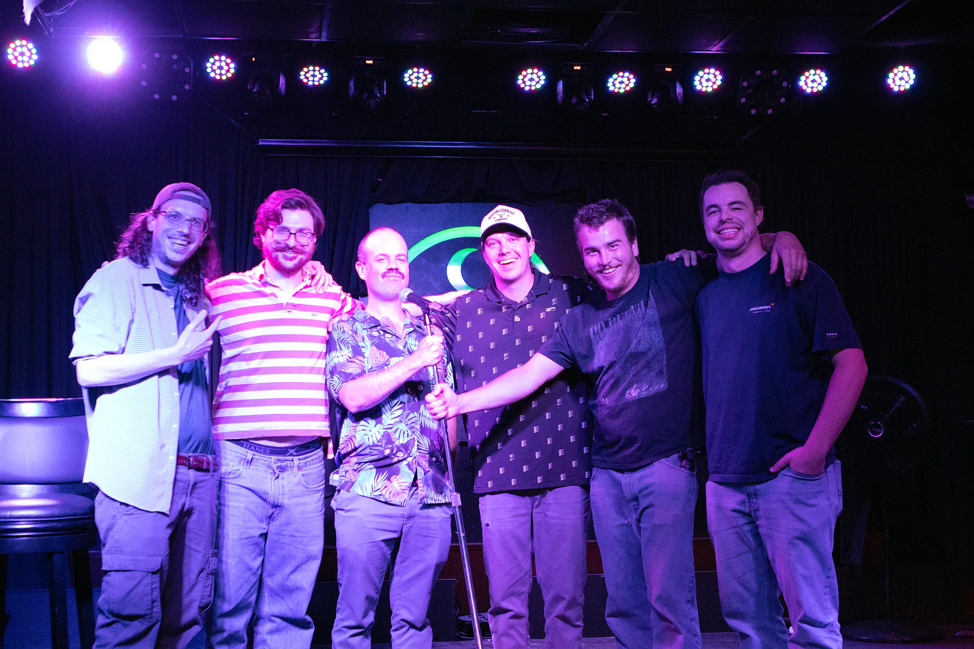 Six comics lined up shoulder to shoulder across the stage after the show, arms around each other, the mic stand still standing in the middle of them.