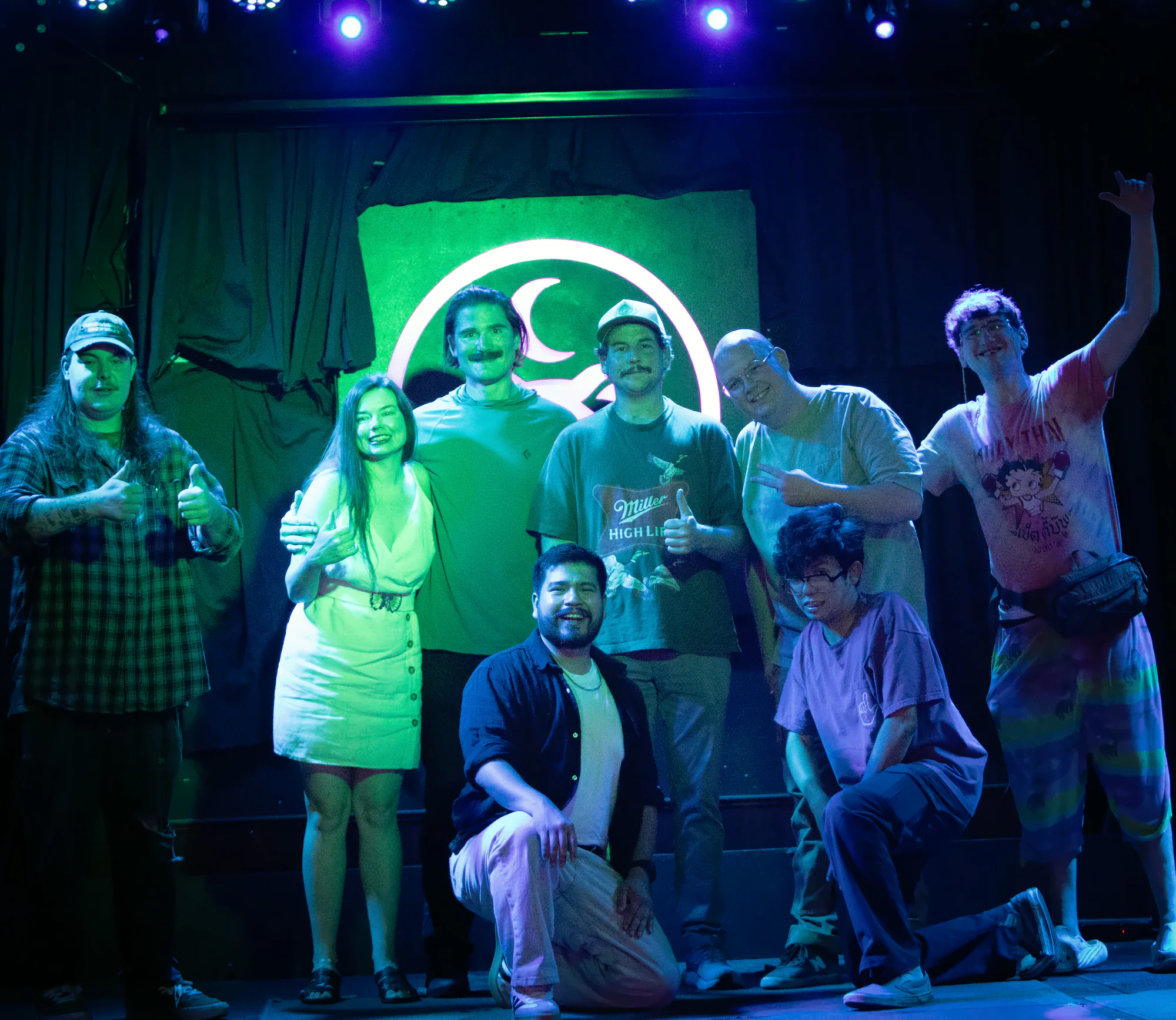 Eight performers posed together on stage, some giving thumbs-up and two crouching at the front, the lit toad’s ring and crescent glowing above their heads.
