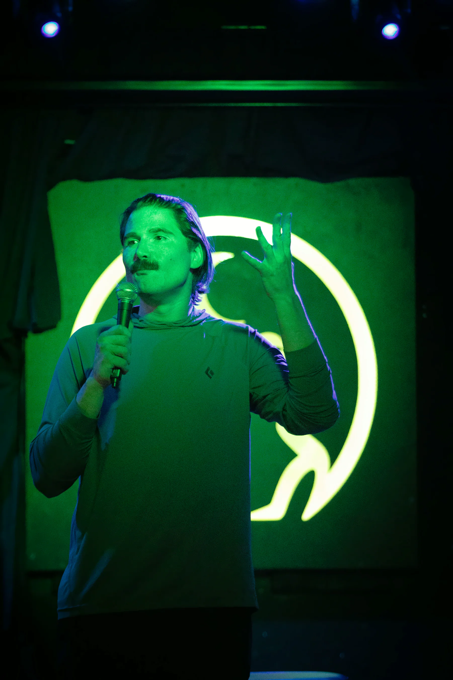 A performer with a moustache in a pale long-sleeved top, one hand raised, the lit toad glowing green behind them with most of the frog hidden.