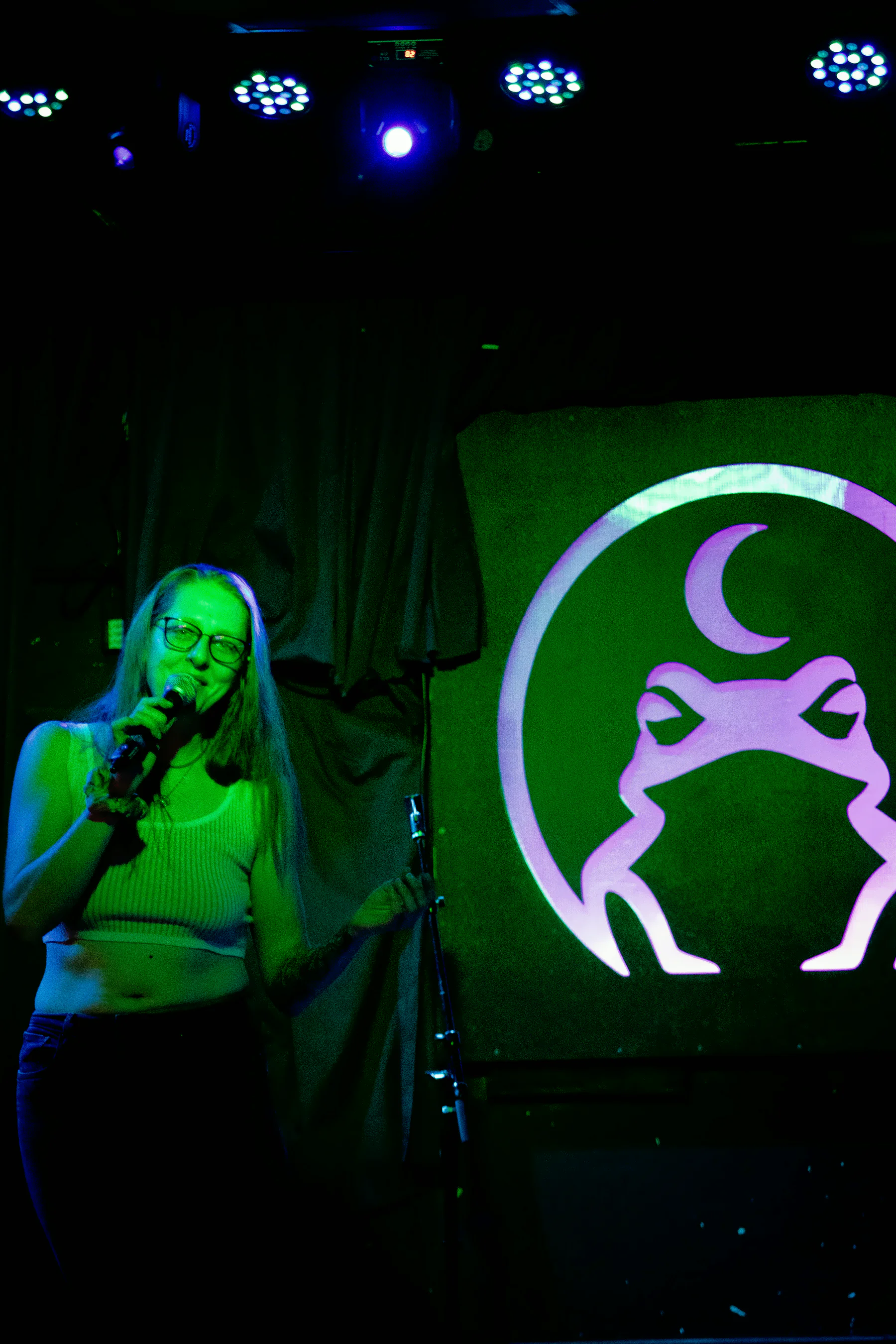 A performer with long hair and glasses at the microphone, the lit toad glowing pale pink on the panel to their right, its far edge cut off by the frame.