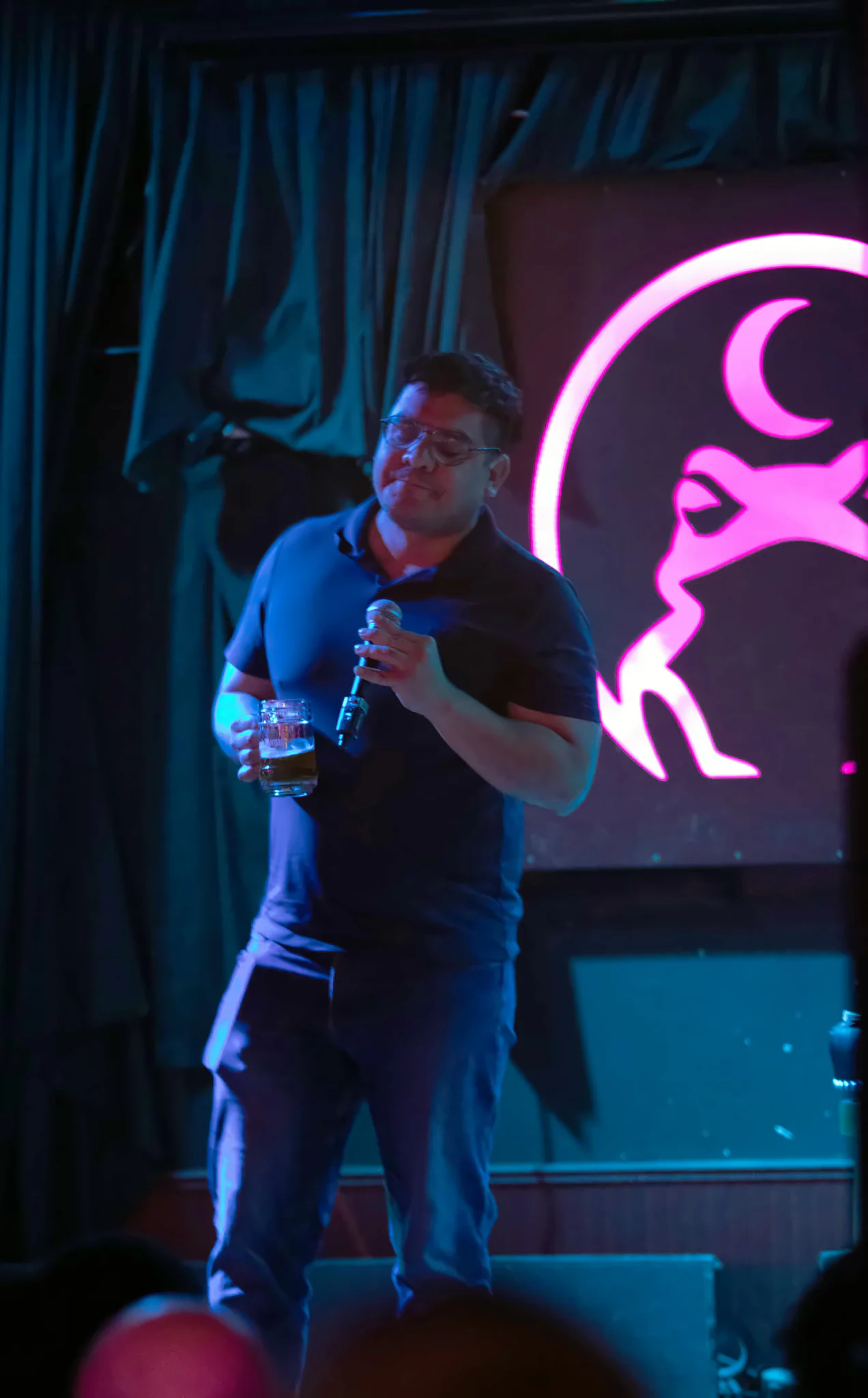 A performer in a navy polo with a microphone in one hand and a glass in the other, the lit toad glowing pink on the wall beside them.
