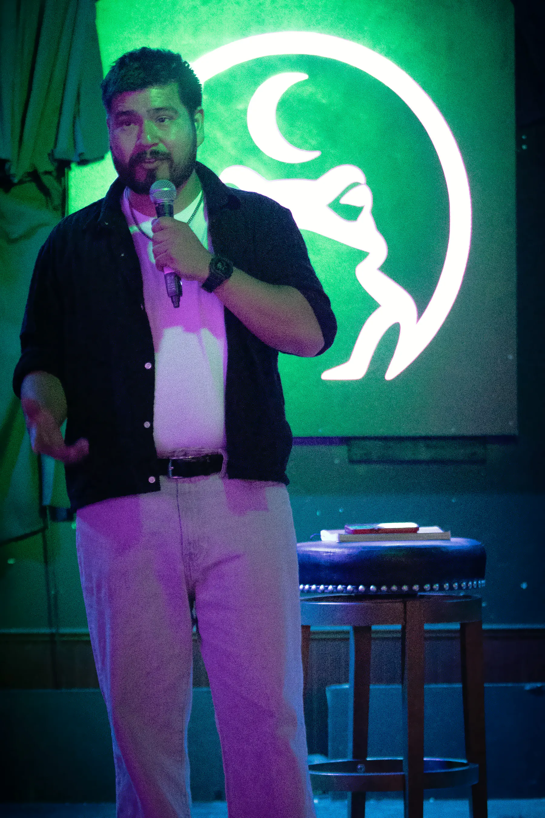 A bearded performer in an open dark shirt at the microphone, the lit toad glowing green on the wall behind them.