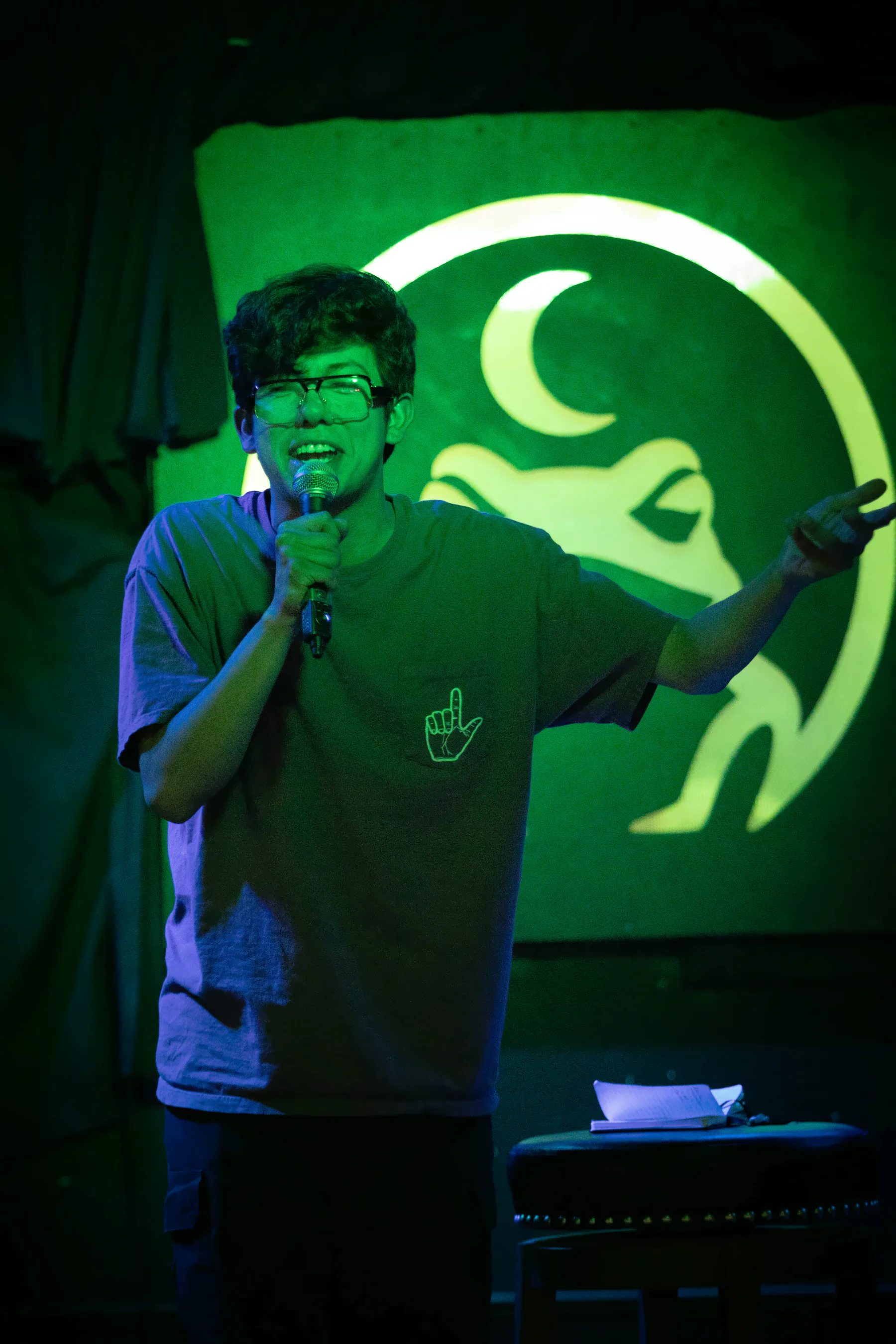 A performer in glasses and a loose tee with a hand-outline print on the pocket, one arm flung wide, the lit toad glowing green behind them.