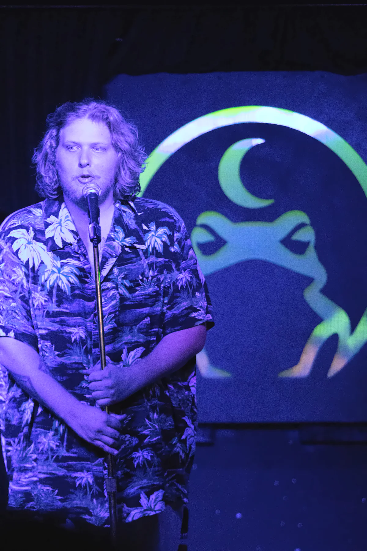 A performer with long curly hair in a palm-print shirt holding a microphone stand, the lit toad glowing green on the wall beside them.