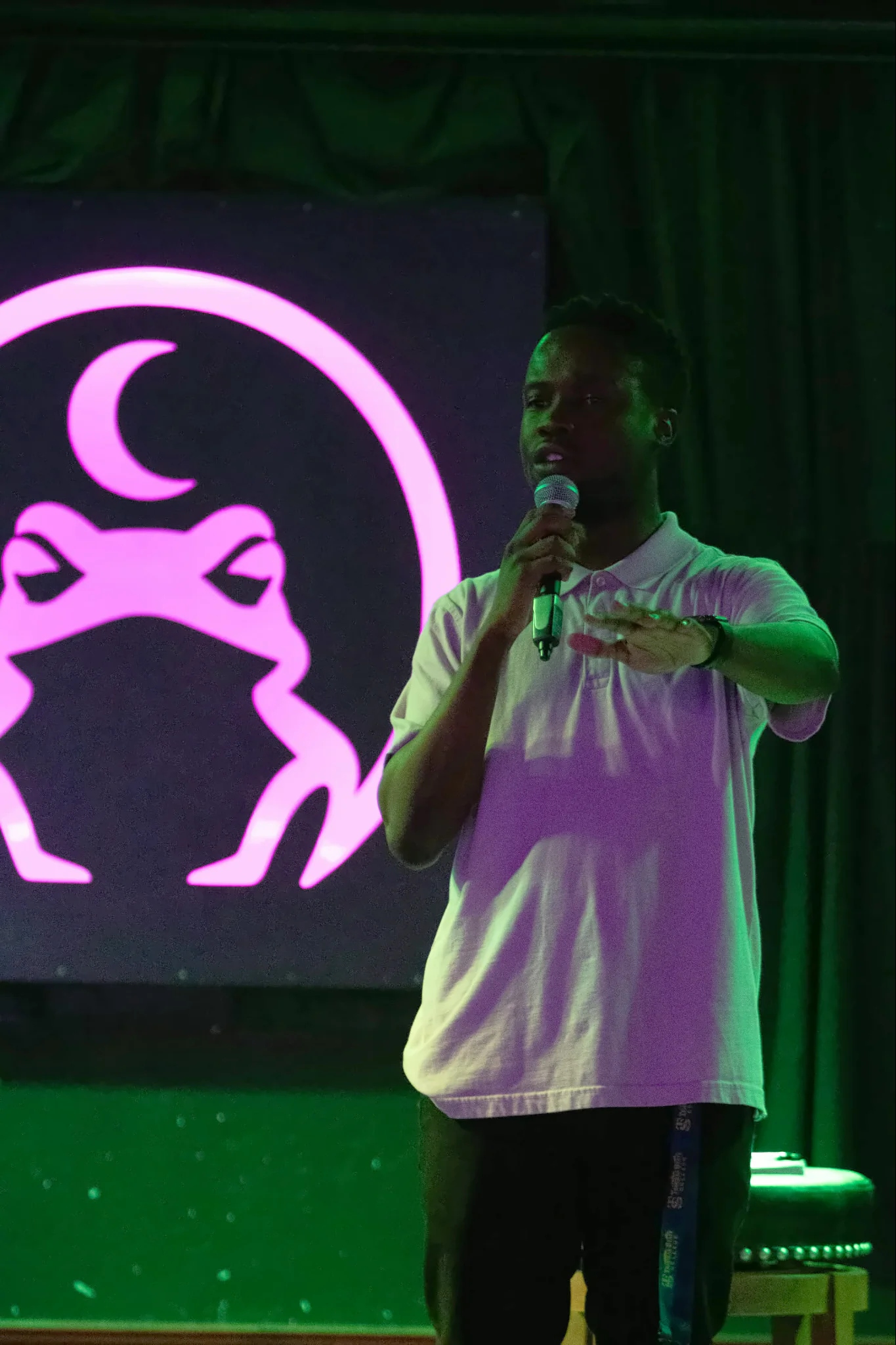 A performer in a pale polo shirt at the microphone with one hand raised, the lit toad glowing pink on the panel beside them.