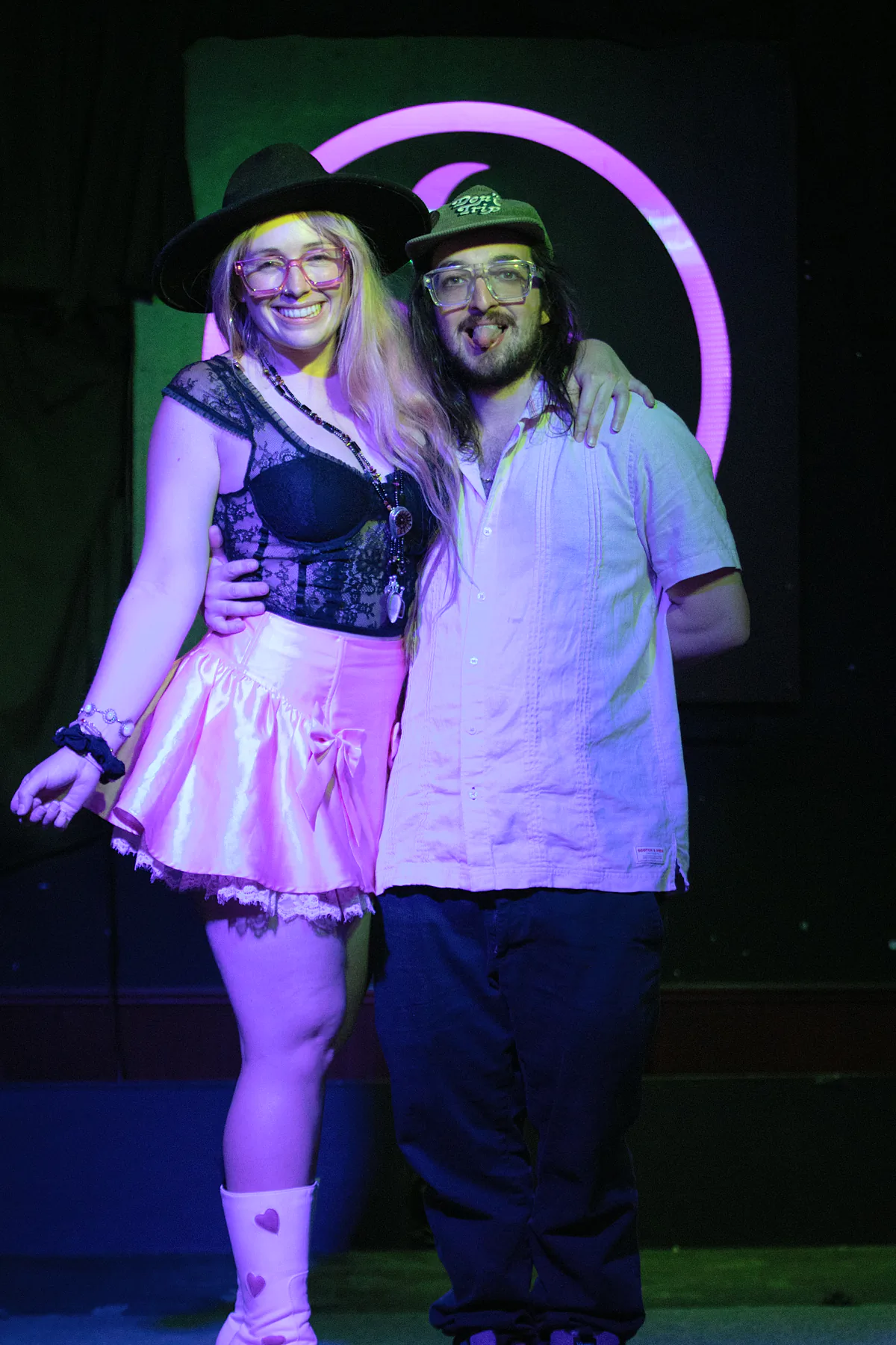 Two performers posing arm in arm at the end of the night, one in a wide-brimmed hat and pink skirt, the other in a cap pulling a face.