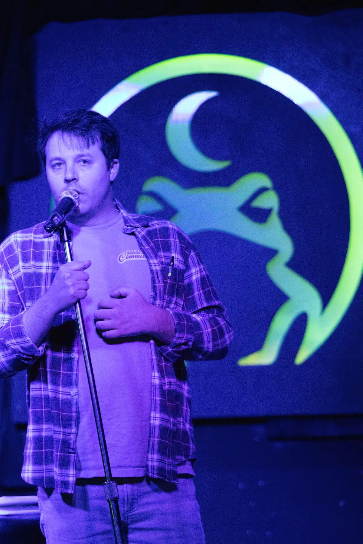 A performer in a plaid shirt over a t-shirt at a microphone stand, the lit toad glowing green on the wall behind them.