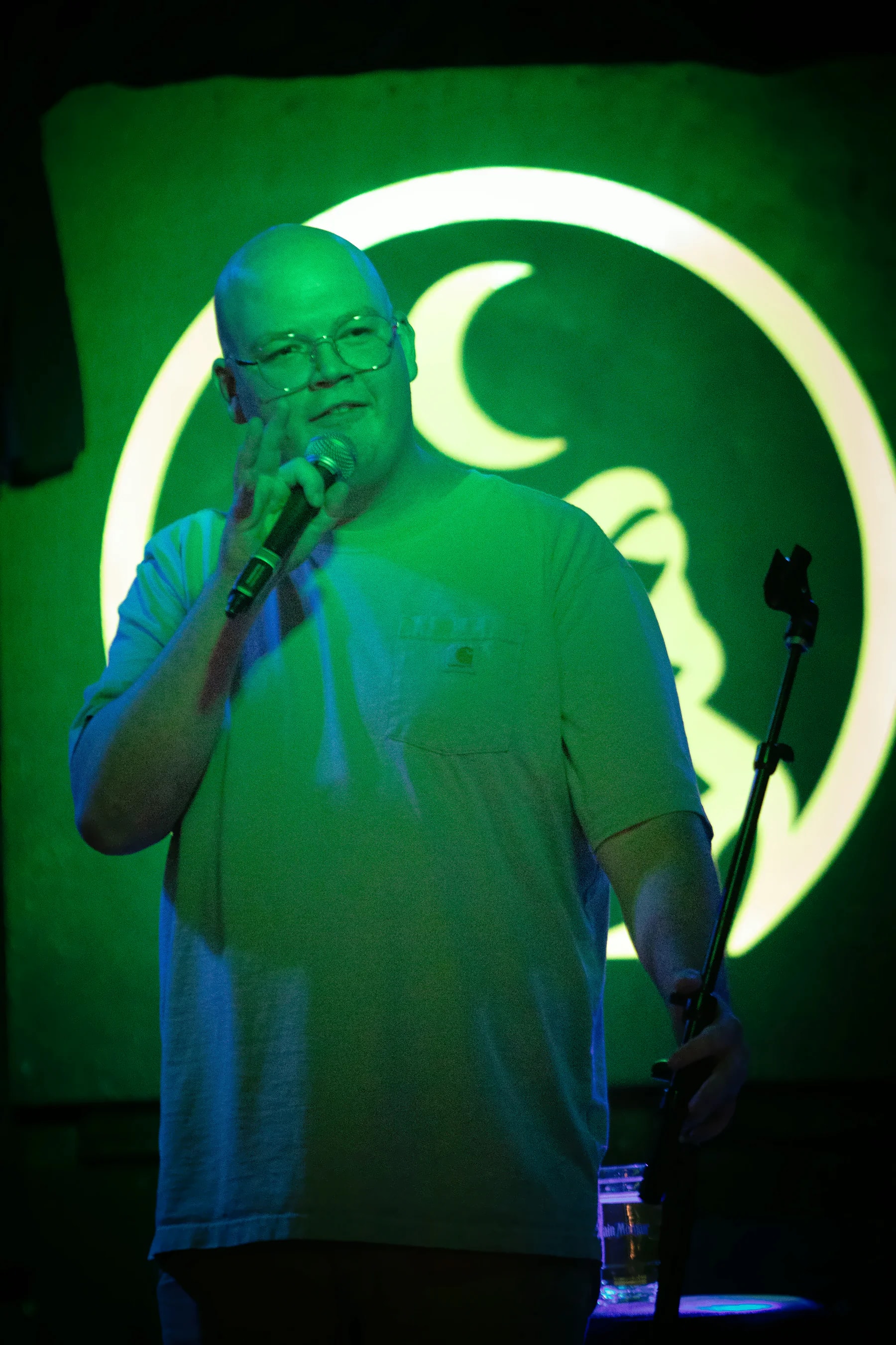 A bald performer in glasses and a pale t-shirt, one hand on the mic stand, the lit toad’s ring and crescent framing their head.