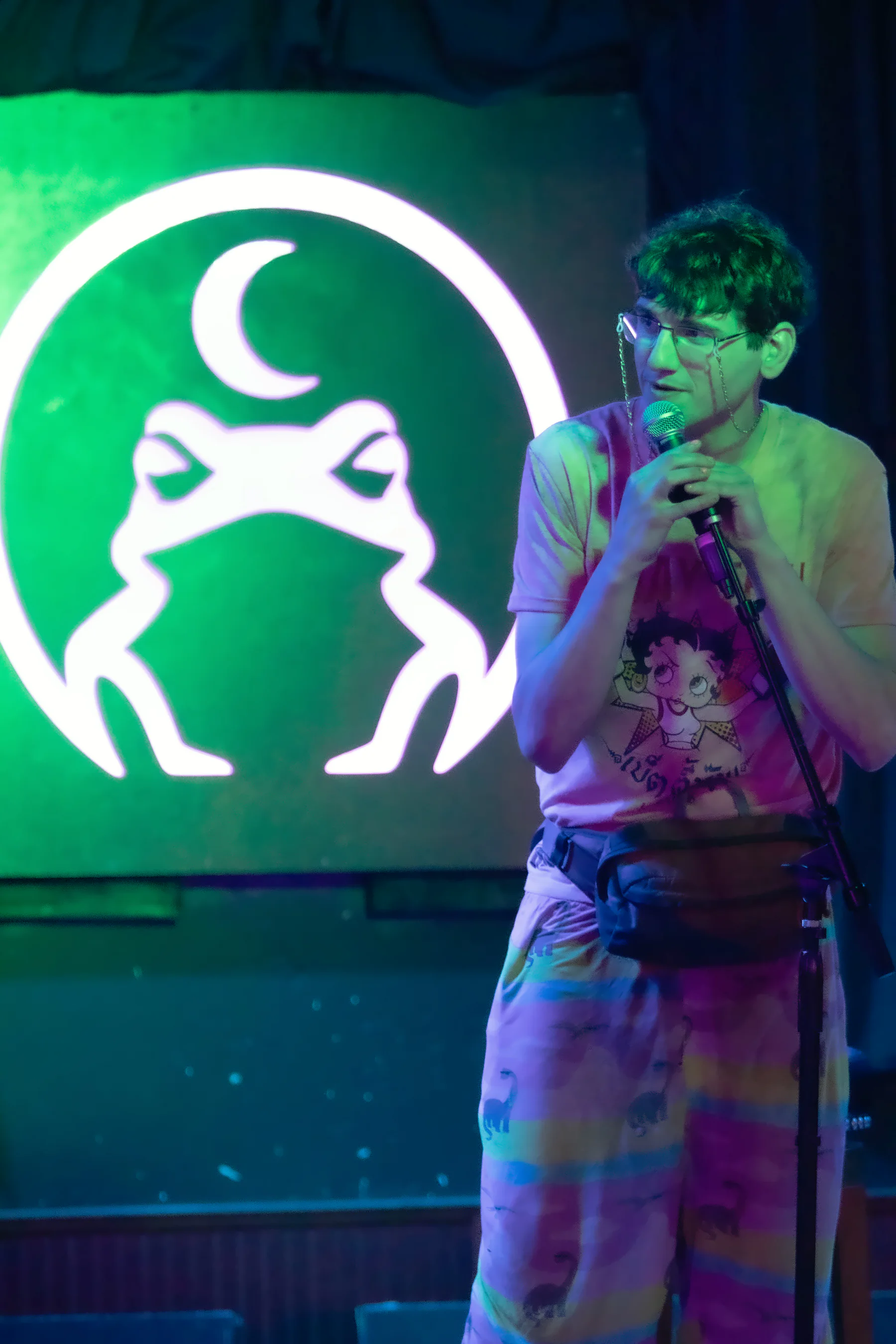 A performer in glasses, a cartoon-print t-shirt and rainbow-striped trousers at the mic stand, the lit toad glowing on the wall beside them.