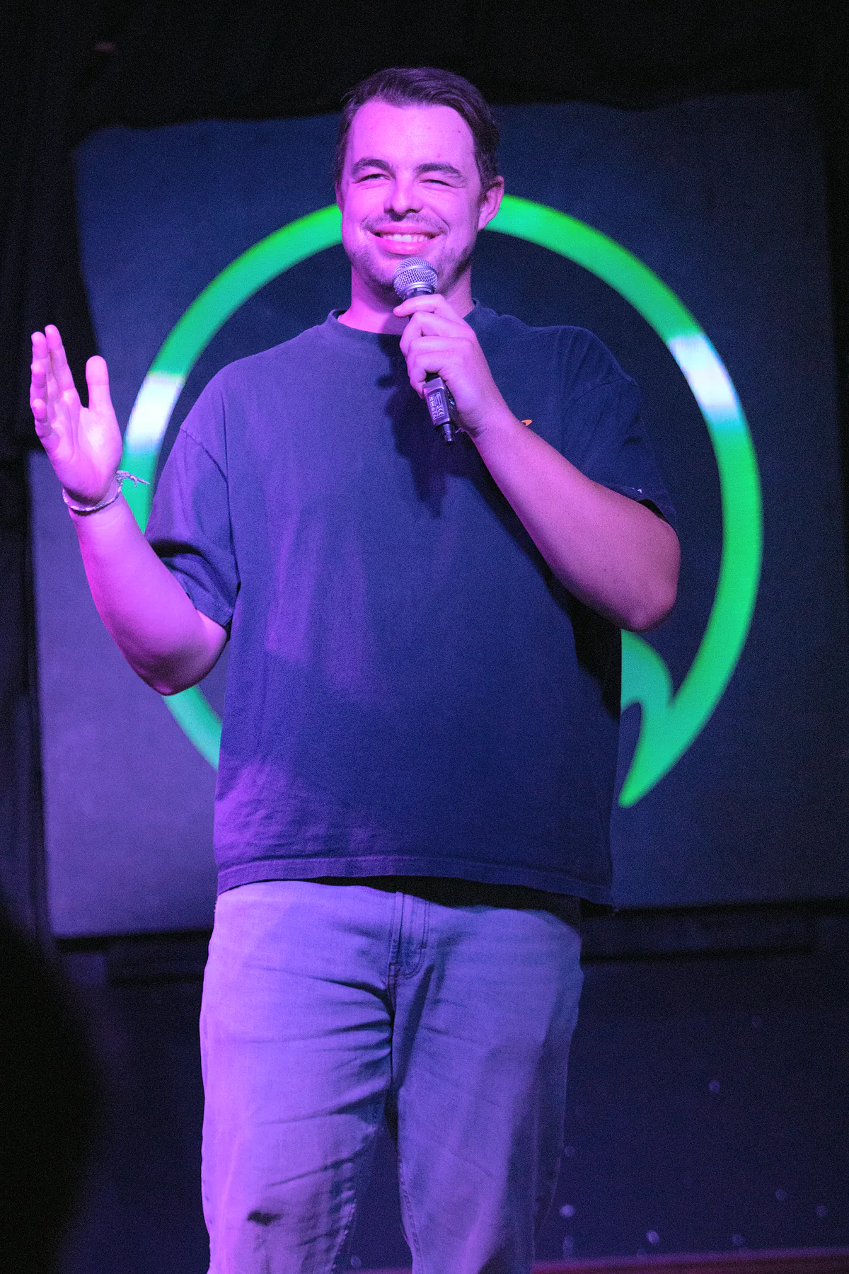 A comic in a navy tee and pale jeans with one hand raised open-palmed, eyes shut, in the middle of a laugh.