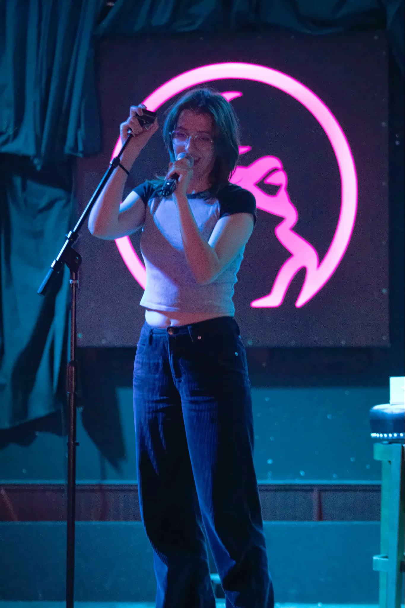 A performer in a white ringer t-shirt singing into a microphone on a stand, the lit toad glowing pink on the wall behind them.