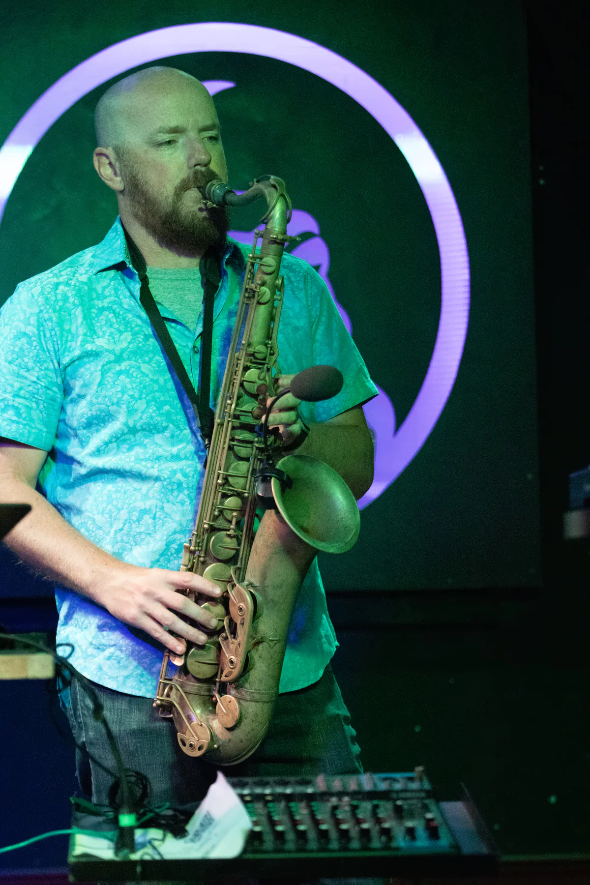 A saxophonist in a patterned short-sleeve shirt playing behind a mixing desk, the lit toad’s ring glowing lavender on the wall behind them.