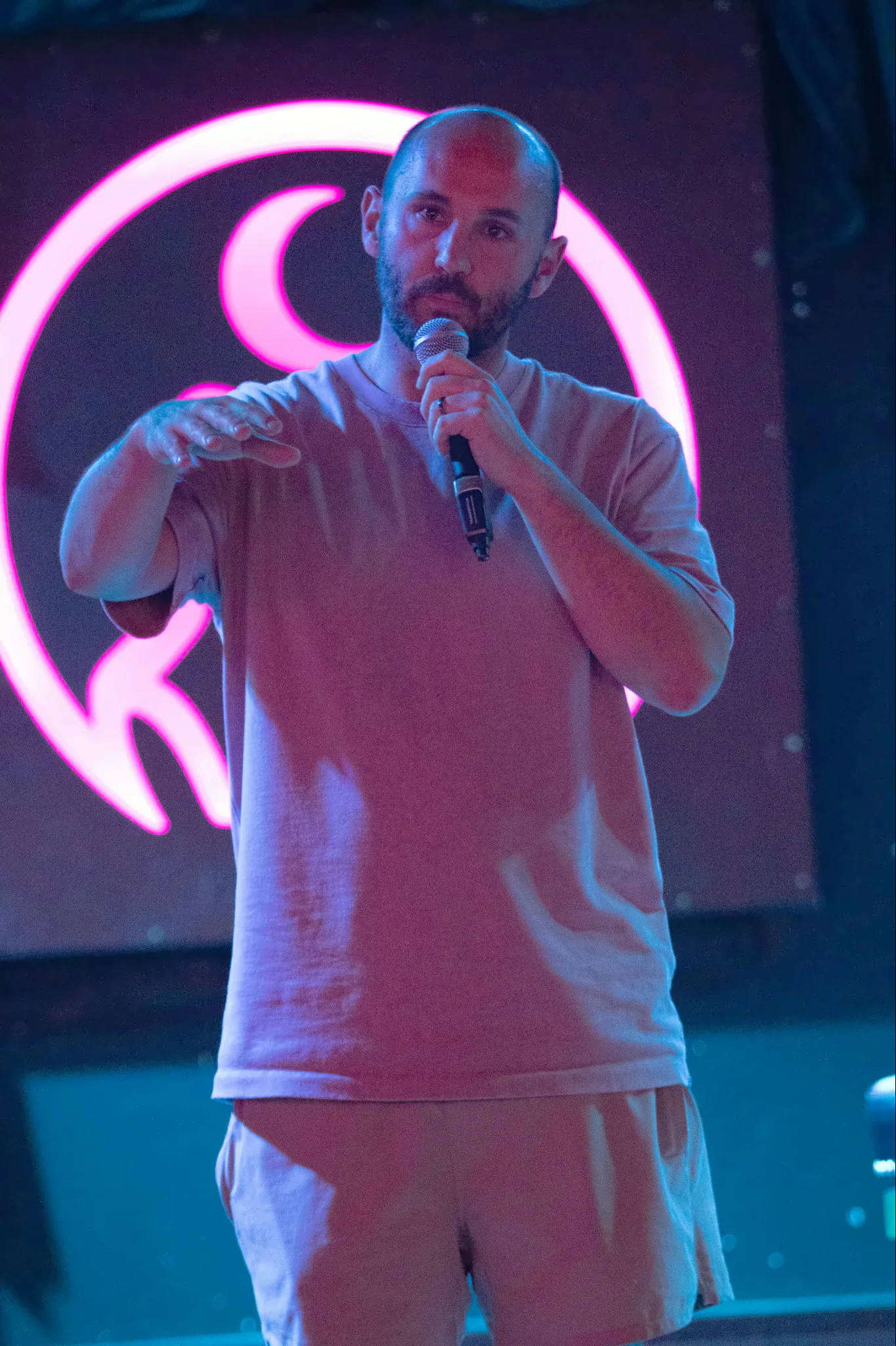 A bearded performer in a pale tee, one hand held flat in front of them, the lit toad’s pink ring showing around their shoulders.