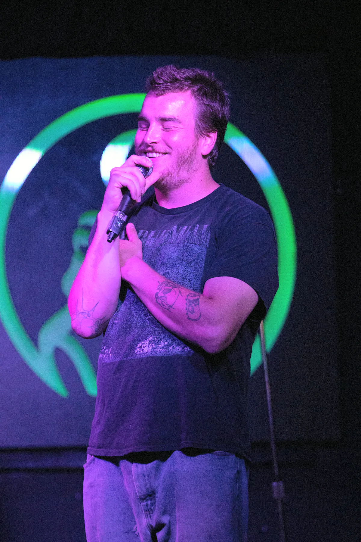 A comic in a faded black band tee with tattooed forearms, holding the microphone close and laughing at his own bit.