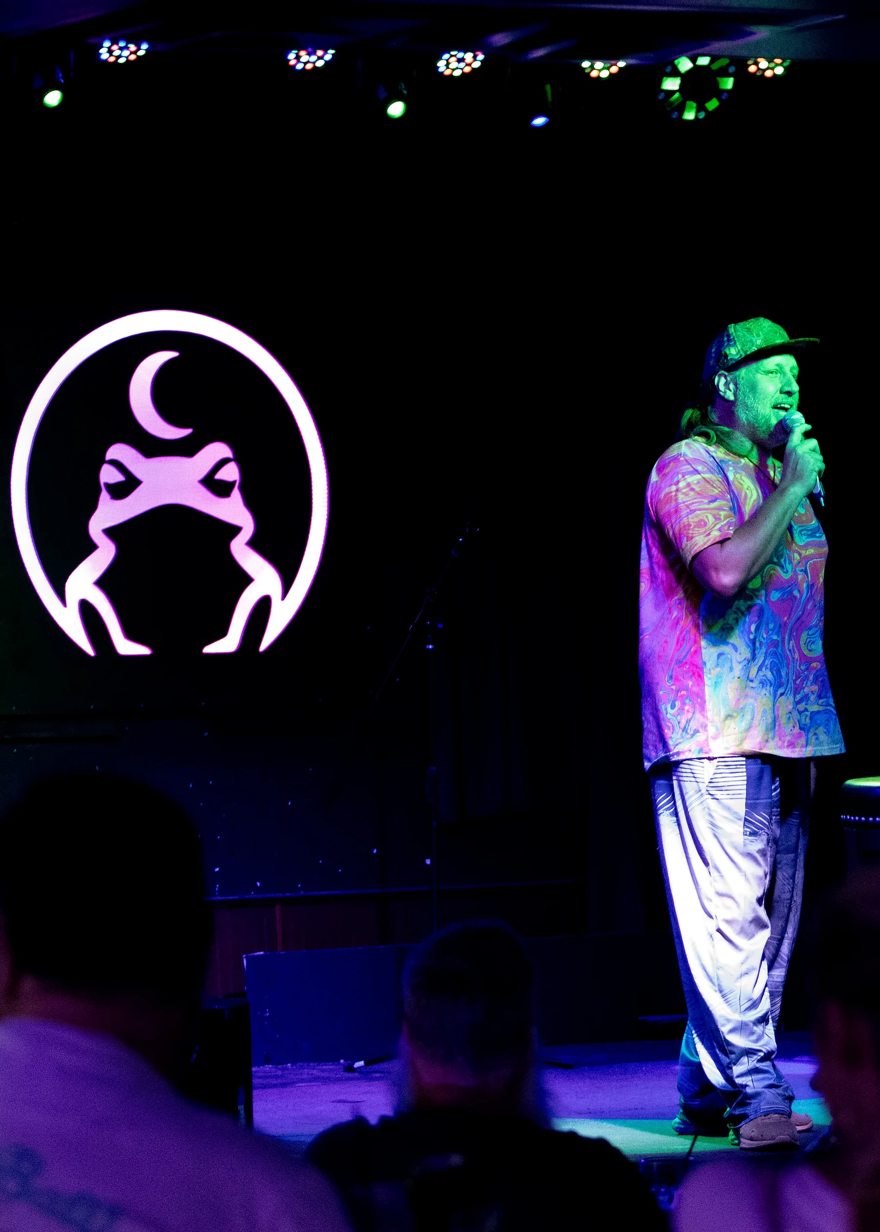A comic in a tie-dye shirt and cap on stage, the heads of the audience silhouetted in the foreground.