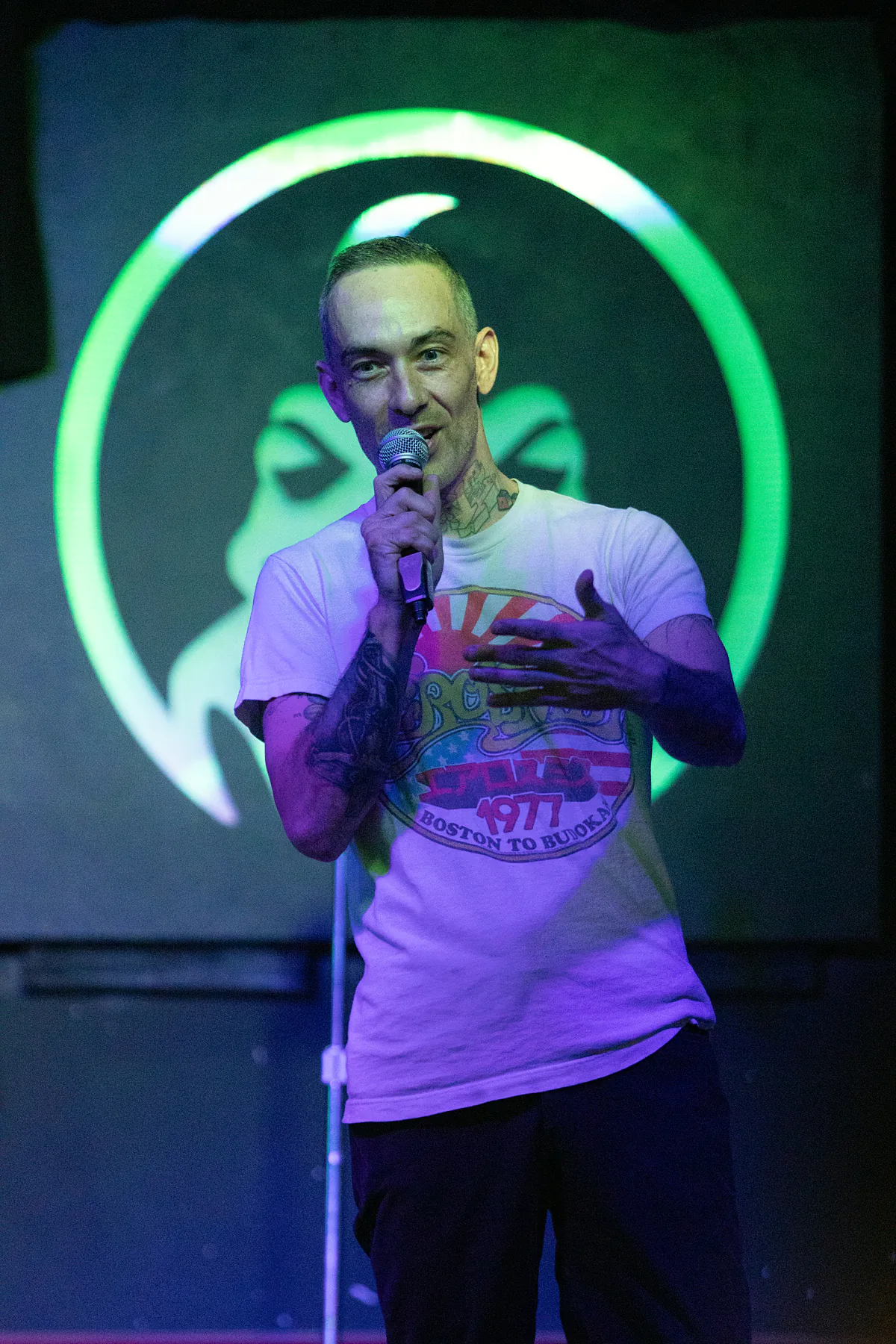 A tattooed comic in a white 1977 tour tee gesturing with an open hand while holding the microphone, lit green from behind.