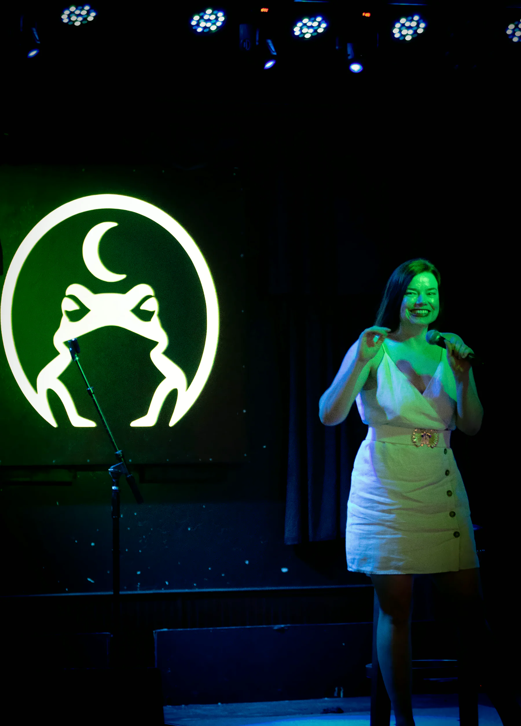 A smiling performer in a light sleeveless dress with a wide belt, microphone in hand, the lit toad glowing on the wall beside them.