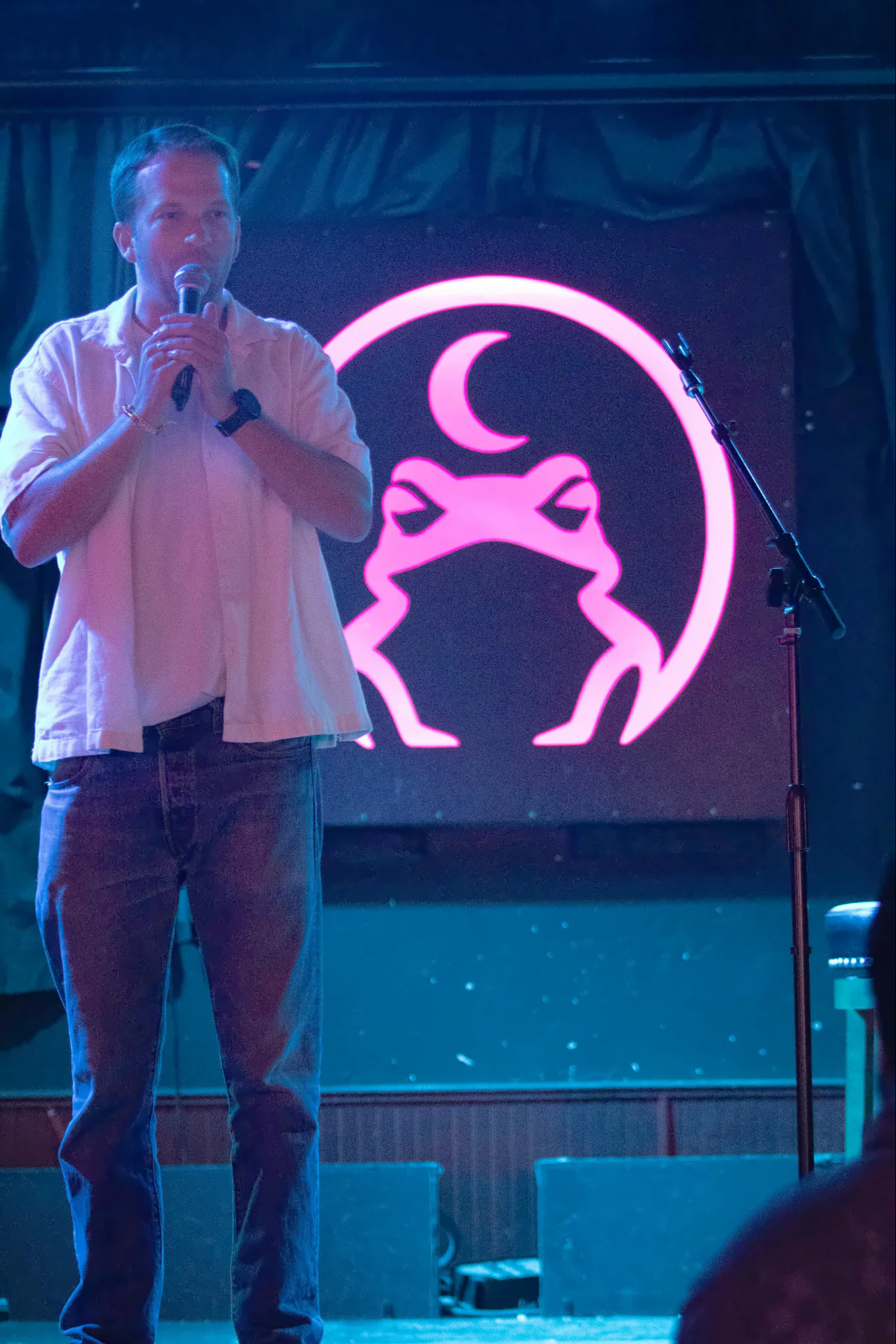 A performer in an open white shirt holding the microphone in both hands, the lit toad glowing pink on the wall behind them.