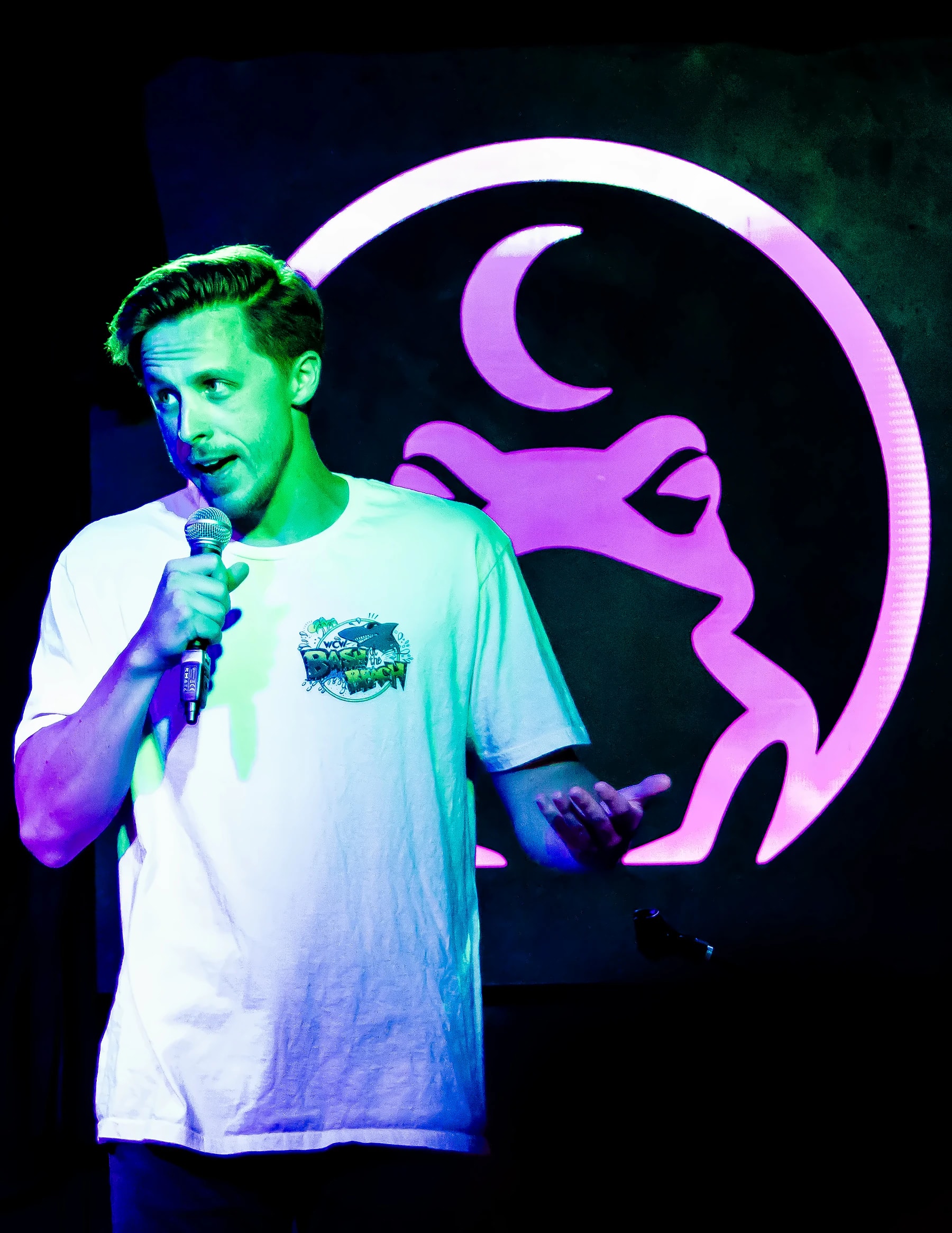 A comic mid-punchline in a white t-shirt, the lit toad filling the wall behind him.